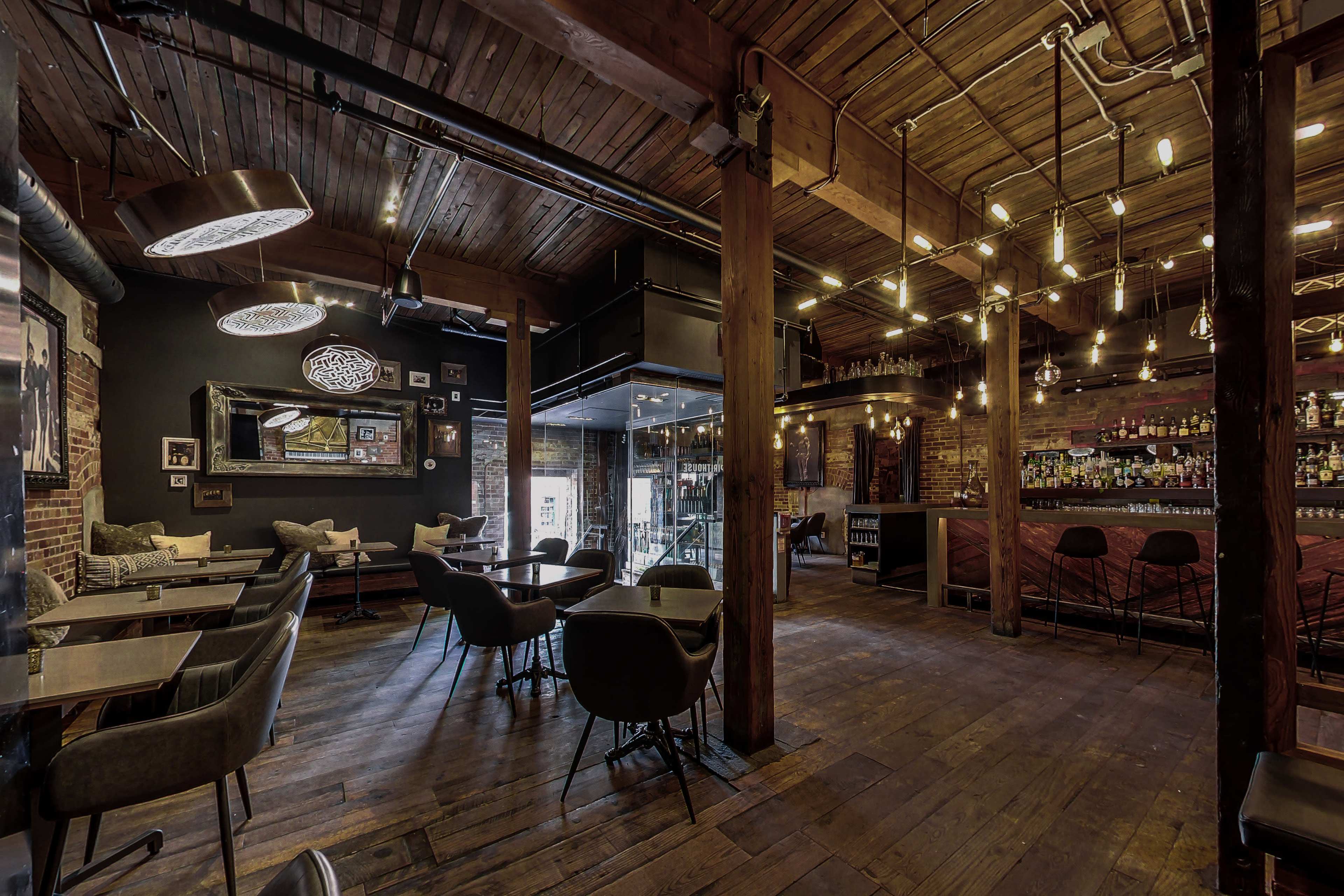 The image shows a rustic bar and dining area with wooden floors, exposed brick walls, and various seating options under warm lighting fixtures.