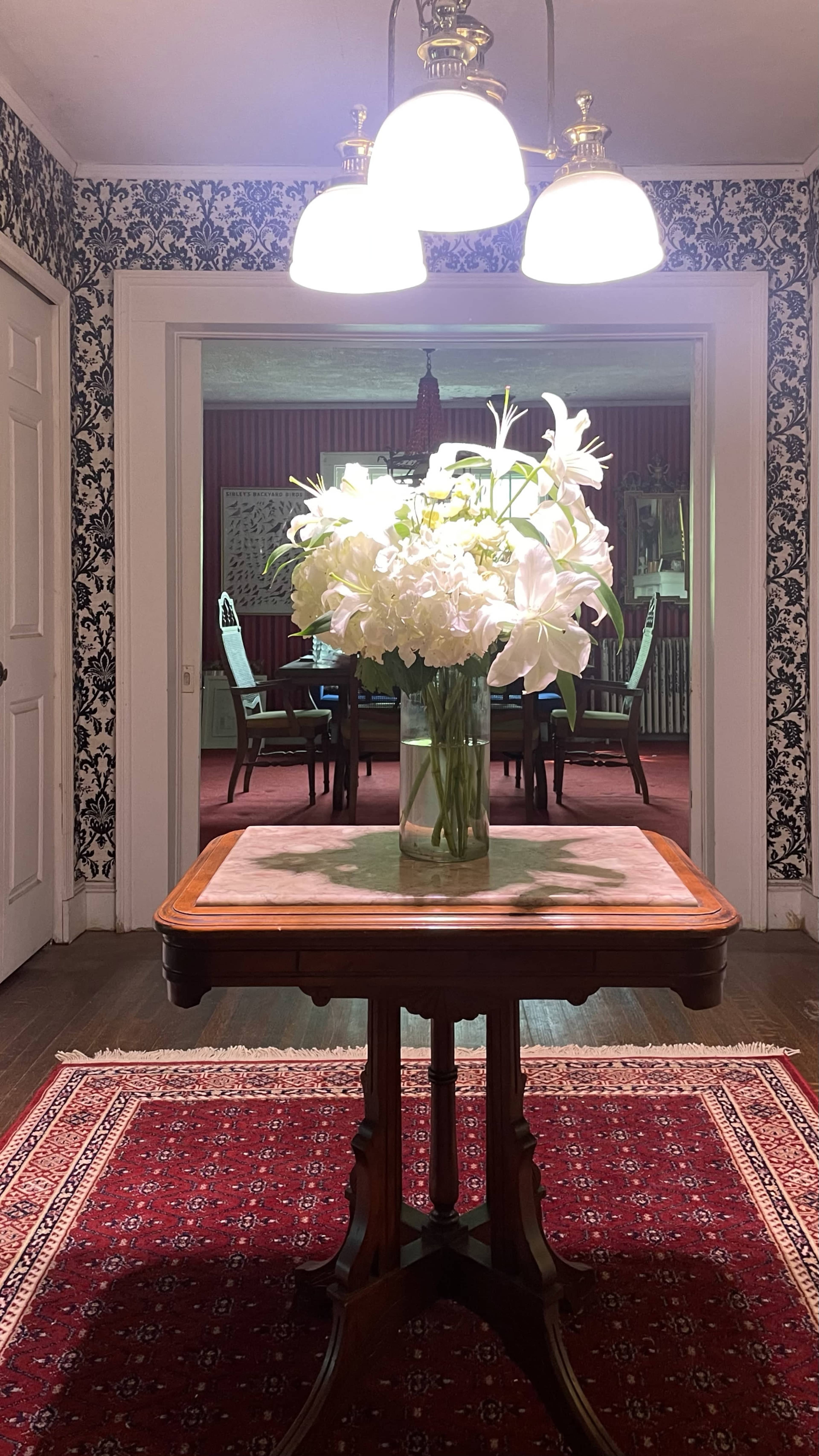 A wooden table with a marble top and a vase of white flowers is positioned in a hallway leading to a room with dining chairs in the background.