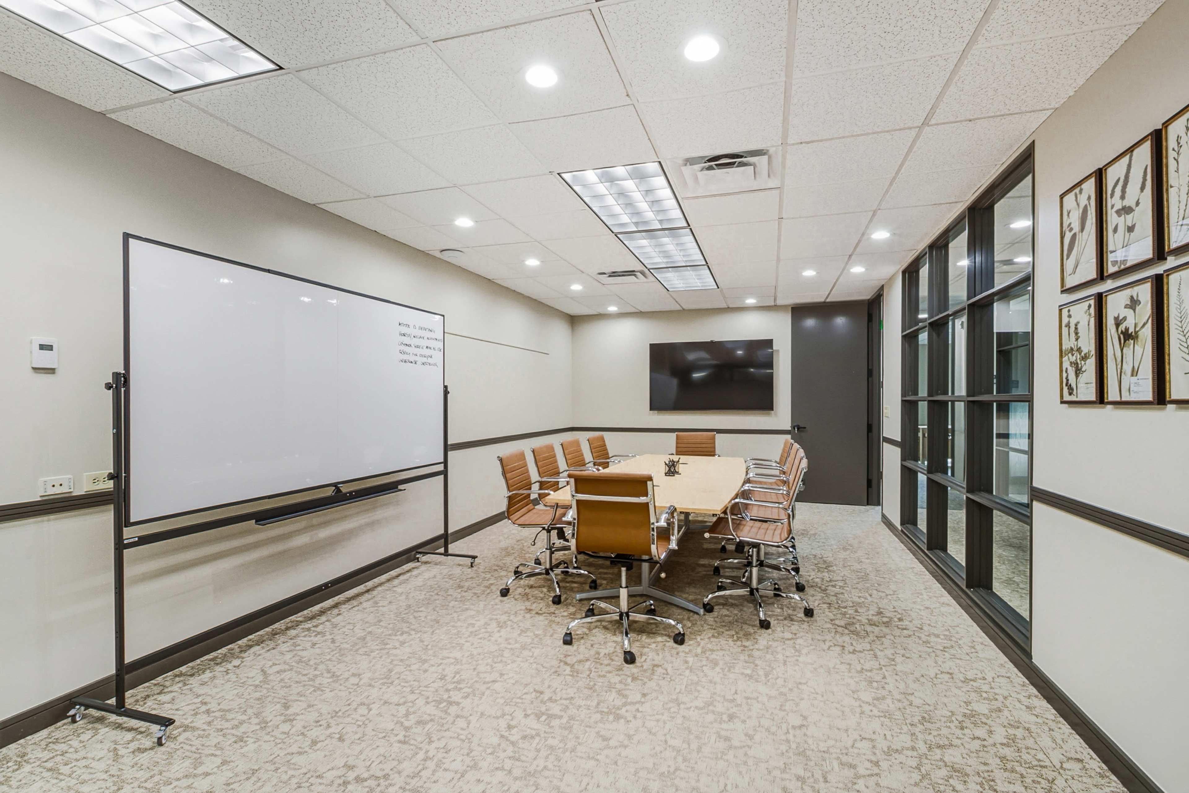 The image shows a modern conference room with a large oval table surrounded by rolling chairs, a whiteboard, a wall-mounted television, and glass-paneled doors.