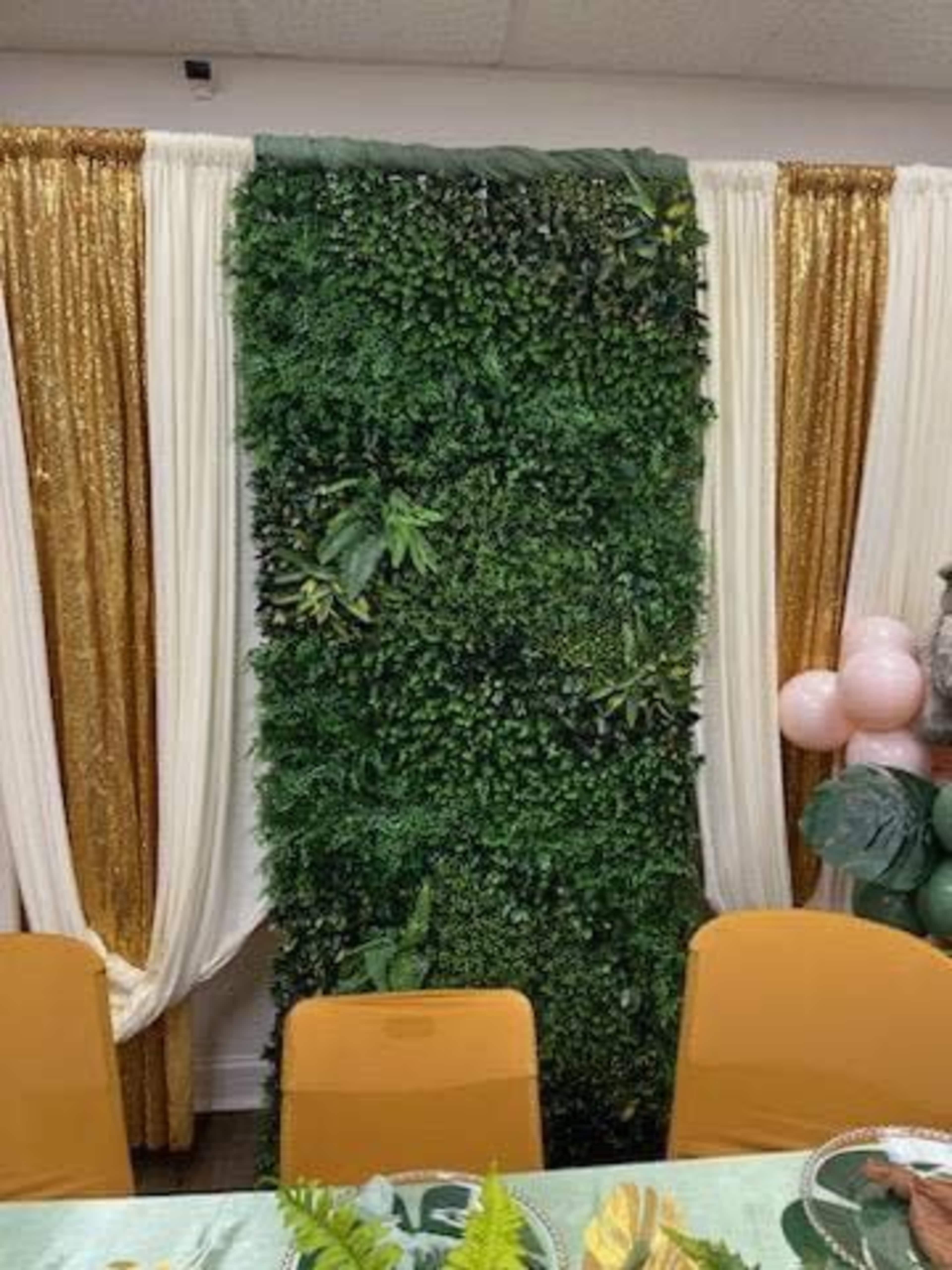 The image shows a decorated backdrop made of lush green foliage, flanked by white and gold drapes in a room setting.