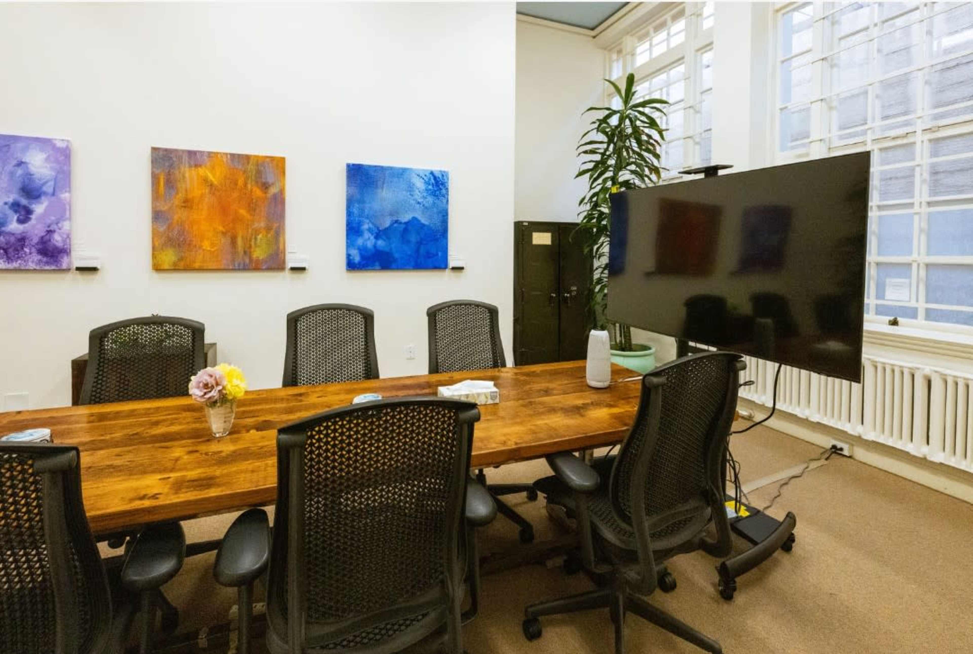 A spacious conference room features a large wooden table surrounded by ergonomic chairs, with colorful abstract paintings on the walls and a large screen at one end.
