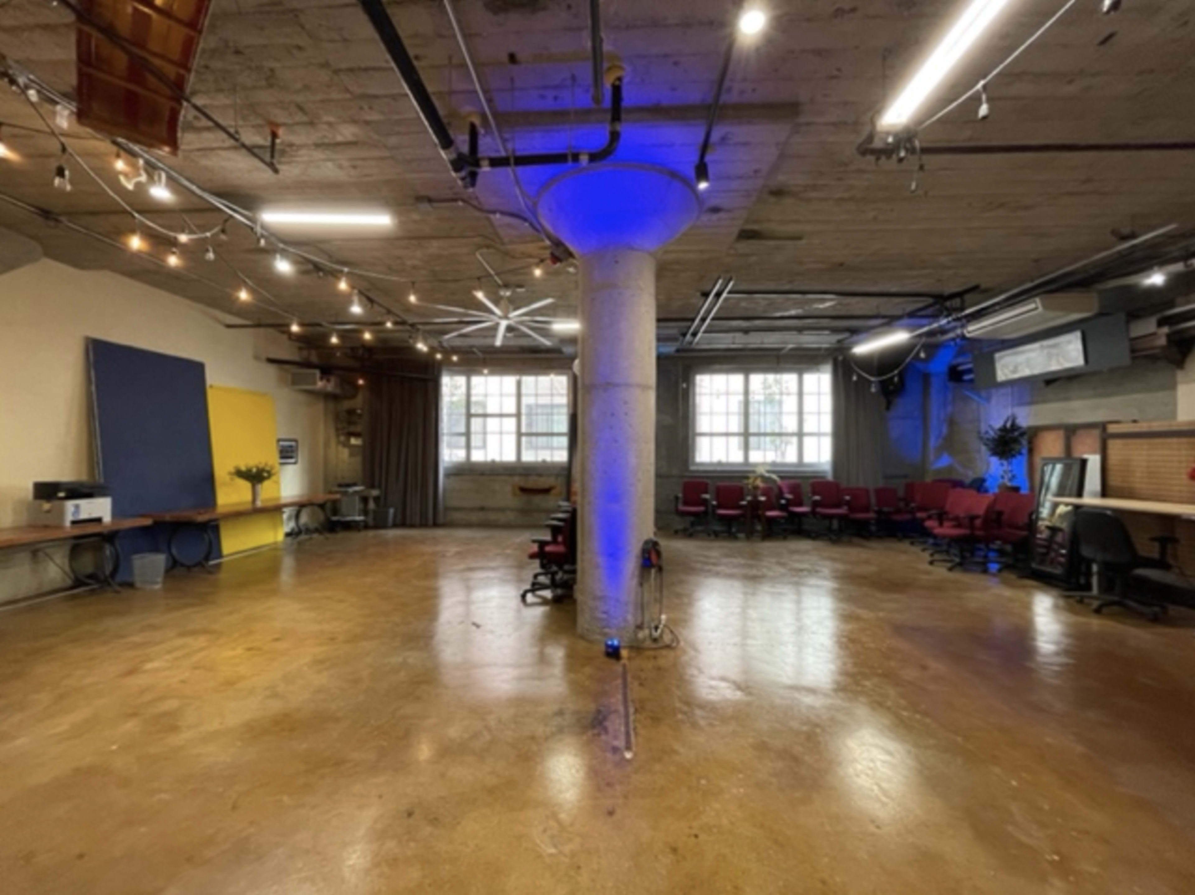 The image shows an industrial-style workspace featuring a central concrete pillar, string lights overhead, and a mix of red chairs and desks against the walls.