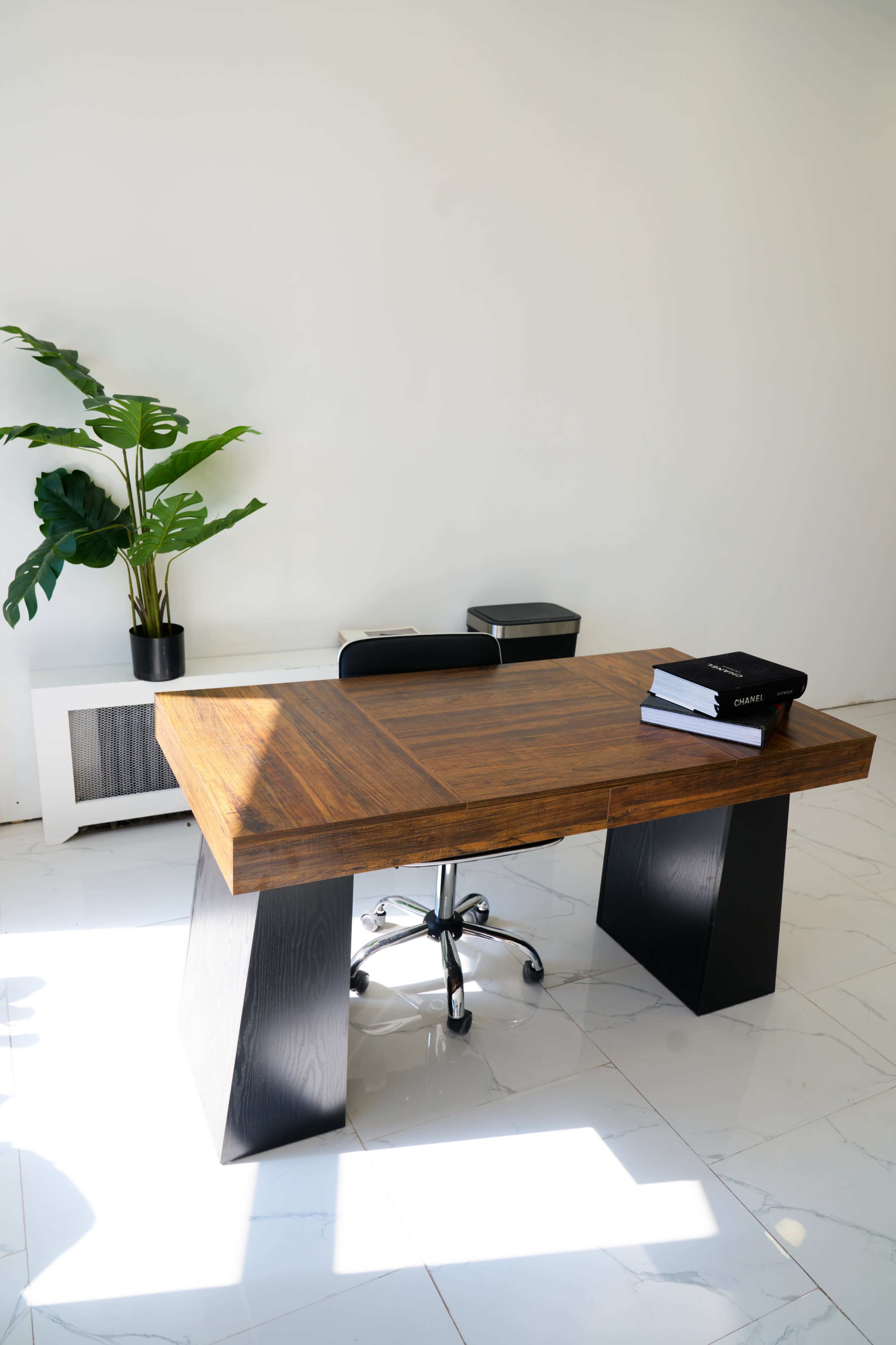 The image shows a wooden desk with a modern design, accompanied by a black office chair, a potted plant, and a white unit in a well-lit room.