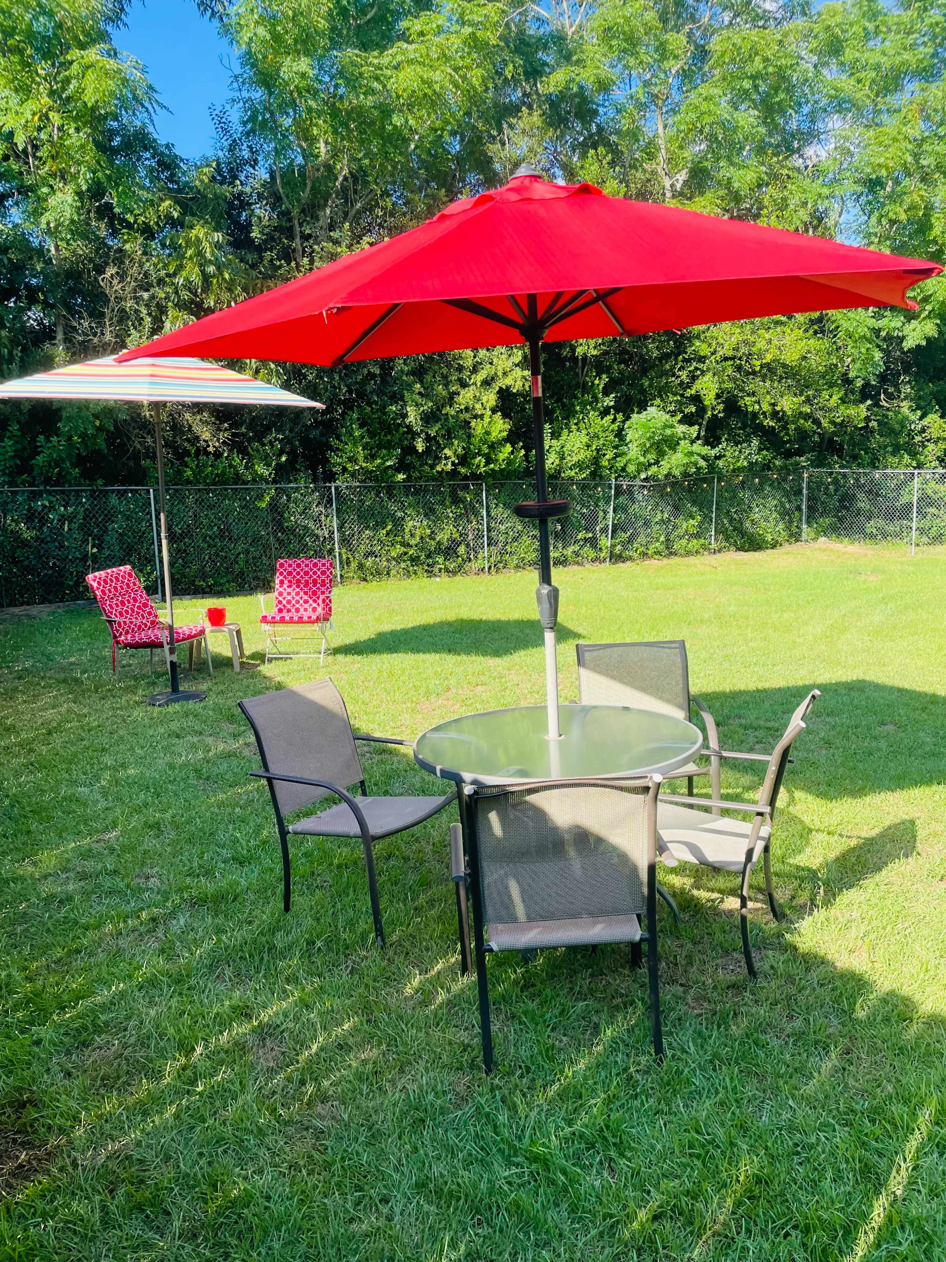 A circular glass table surrounded by six chairs is situated under red umbrellas on a grassy lawn.