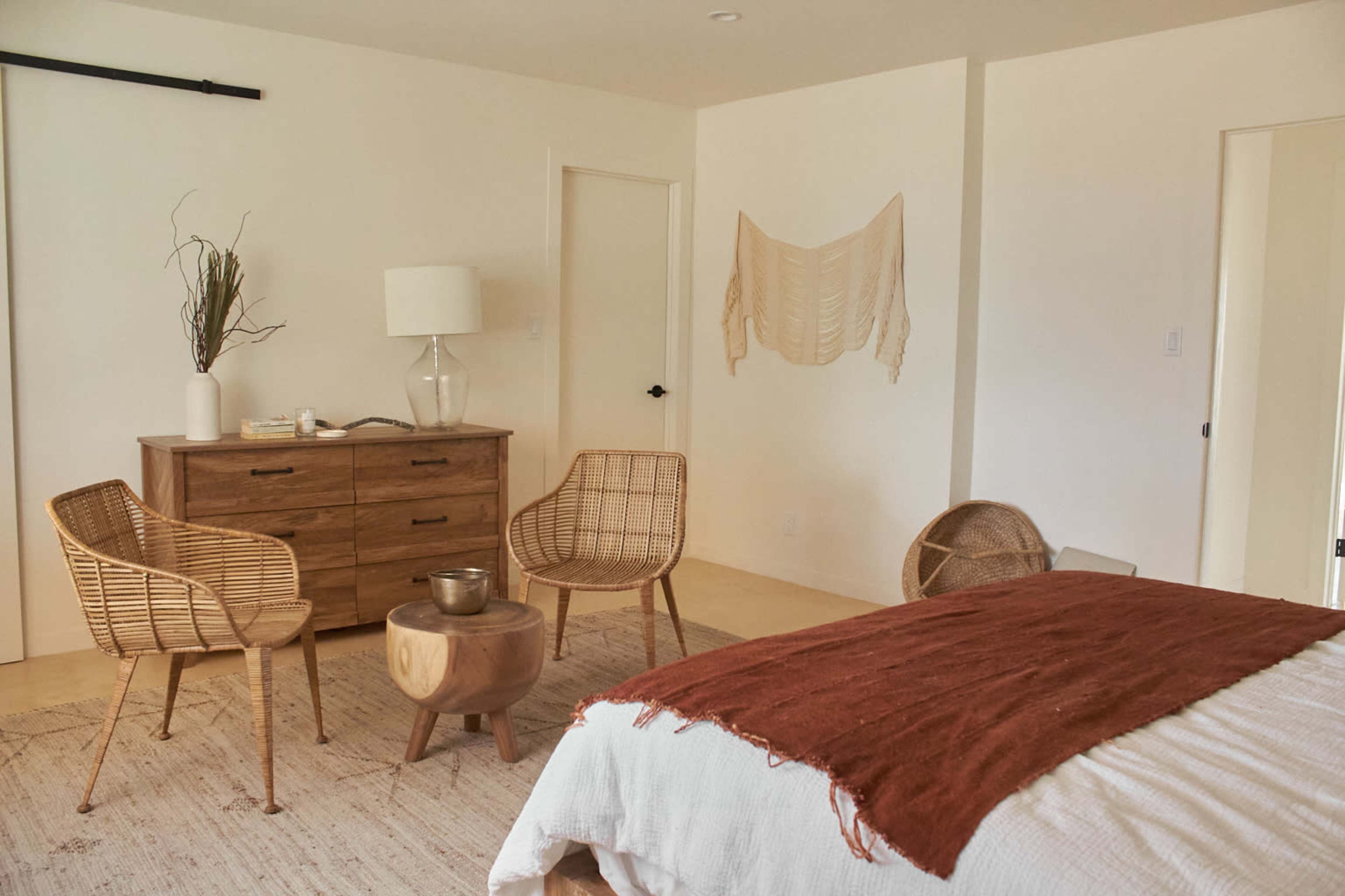The image features a minimalist bedroom with a wooden bed, a woven rug, and a seating area consisting of four wicker chairs and a round wooden table.