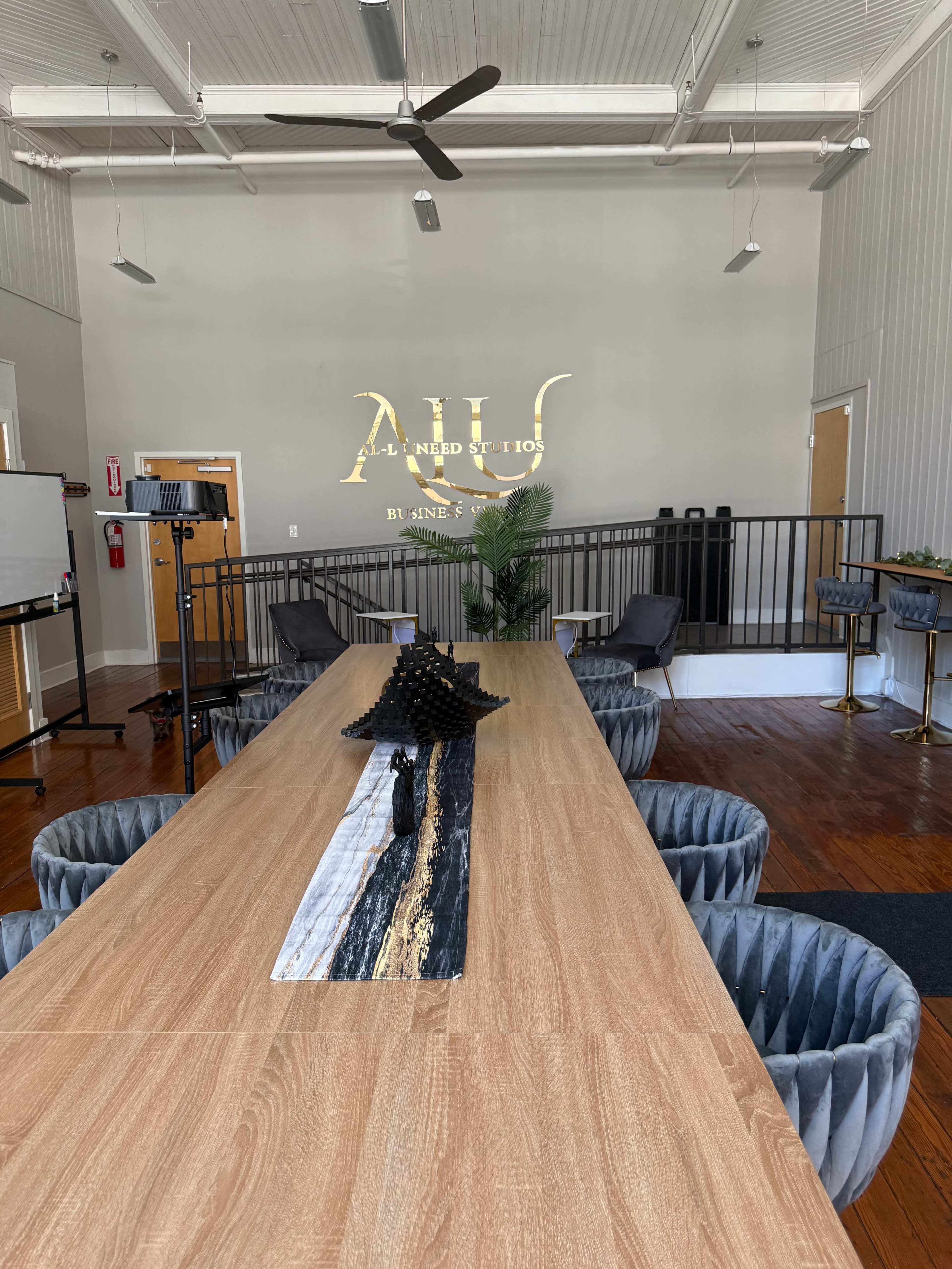 The image shows a modern conference room with a long wooden table, surrounded by circular gray chairs, and featuring a decorative centerpiece and a wall with a logo.