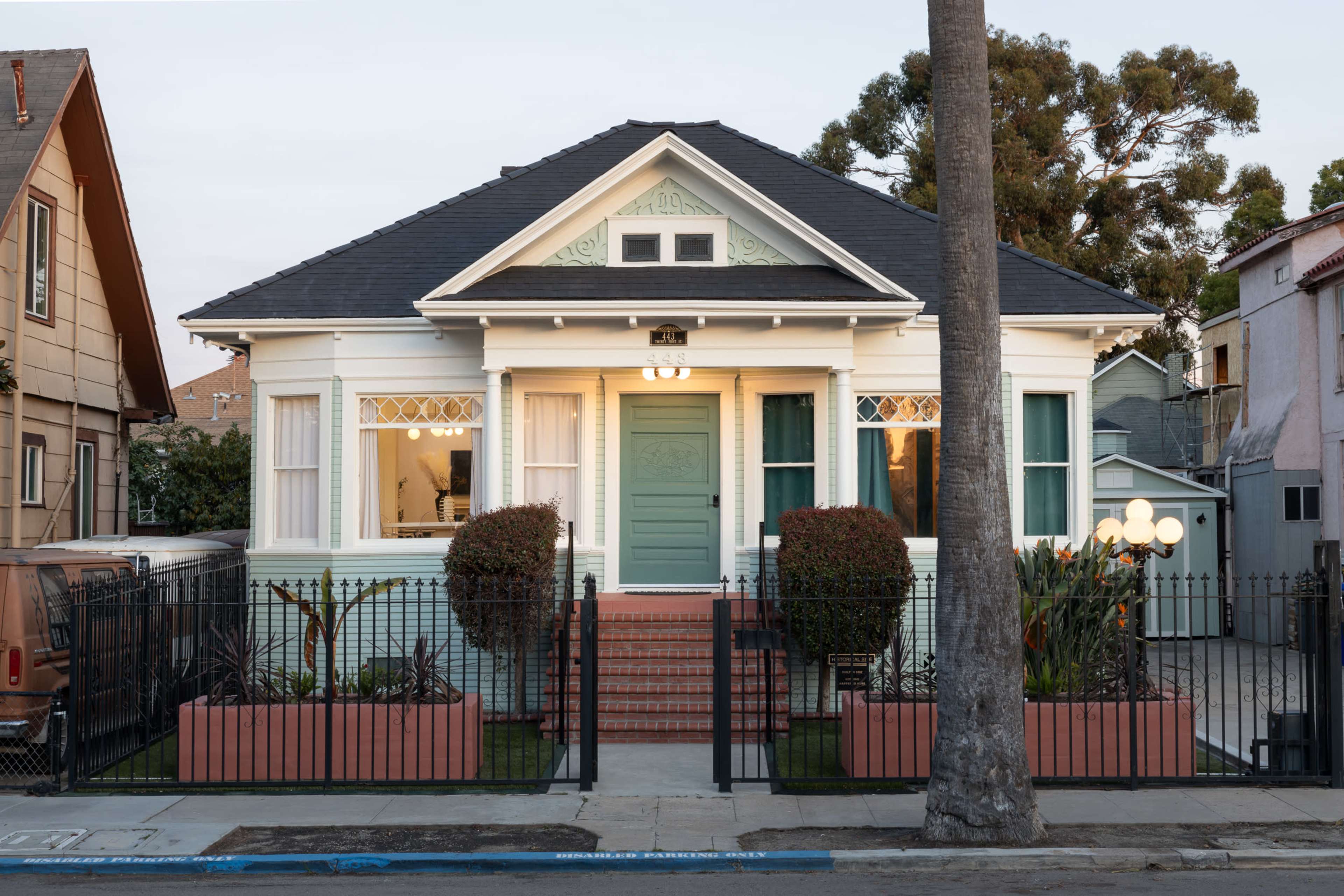 The image shows a single-story house with a light blue door, surrounded by a black fence and neatly arranged landscaping, including two bushy plants in front.