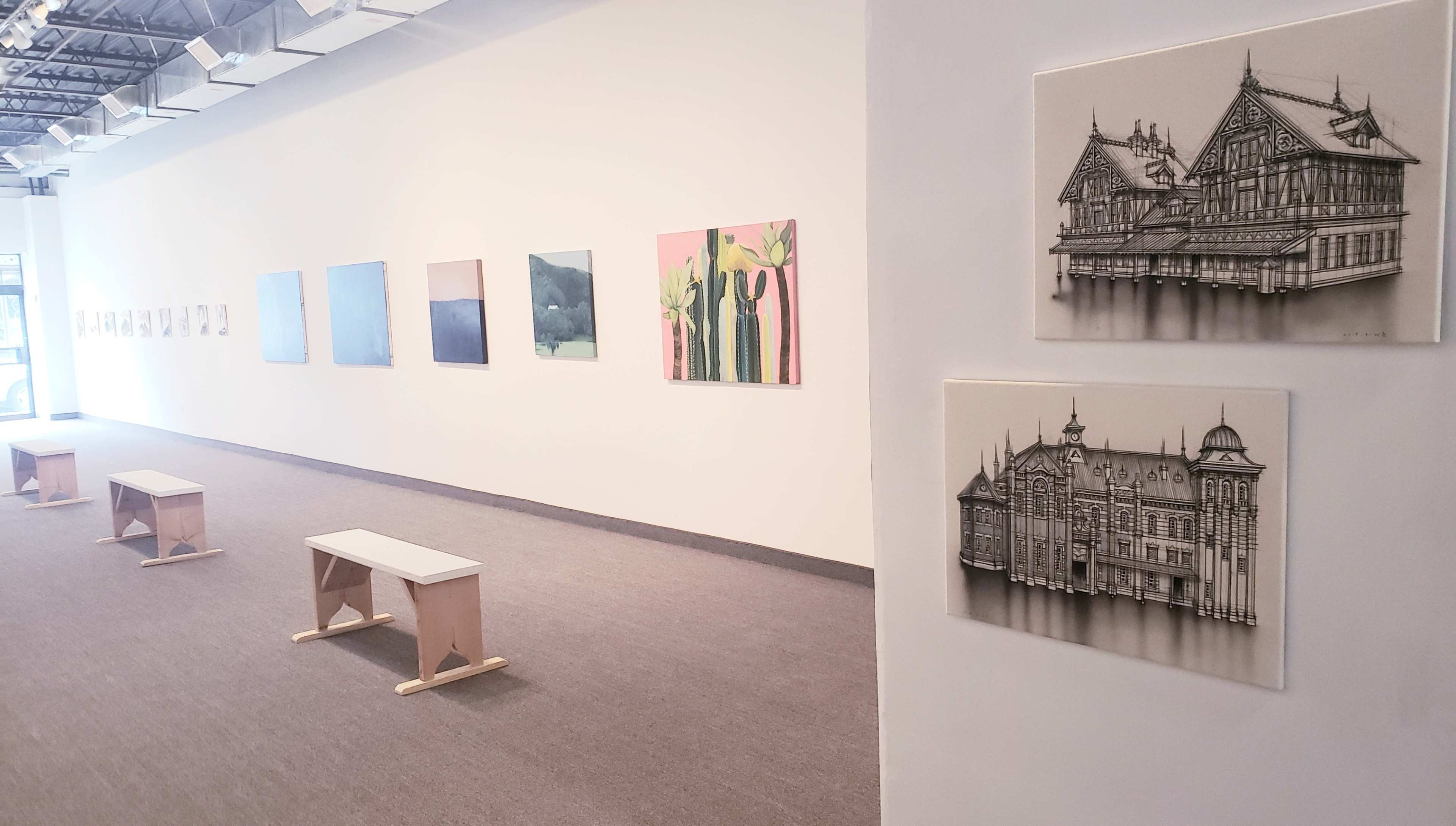 An art gallery with a series of light-colored paintings on the wall and two detailed architectural drawings displayed on the right, along with simple wooden benches arranged in the center.