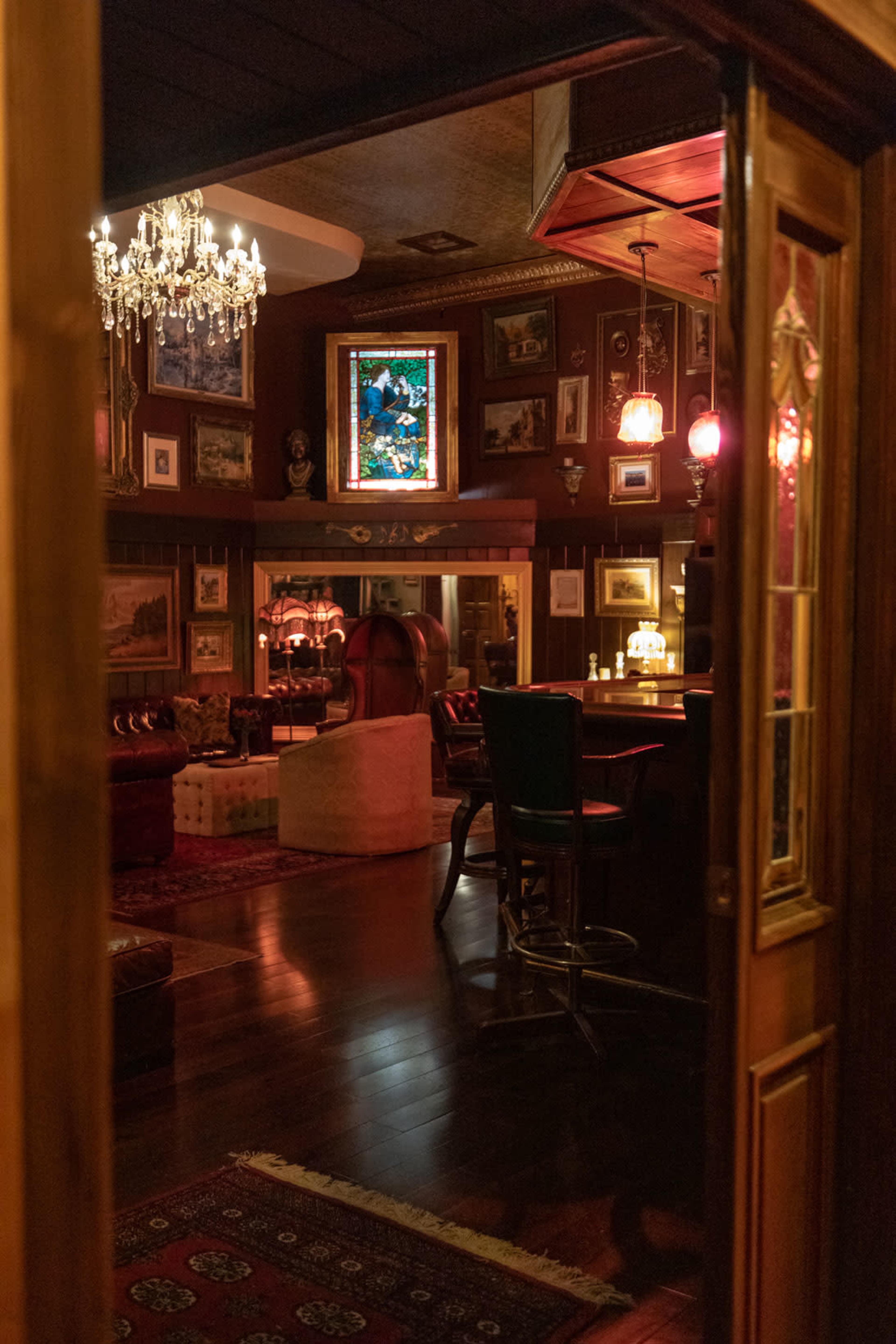 The image shows a dimly lit room adorned with antique-style decor, featuring a chandelier, framed art, and a bar area, with rich wooden finishes and a patterned rug.