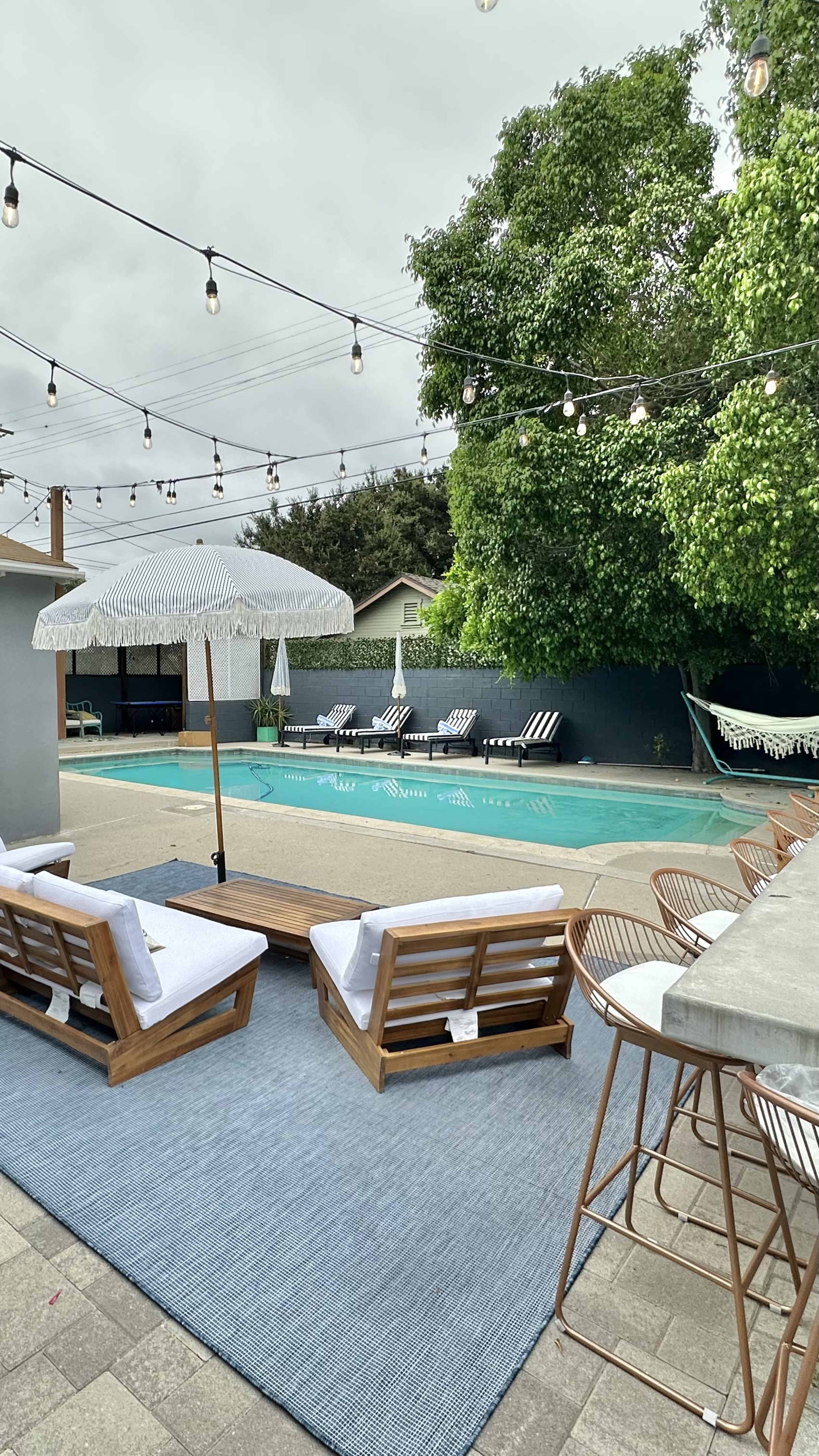 The image shows a backyard pool area with lounge chairs, a large umbrella, and a bar seating arrangement, surrounded by string lights and lush greenery.