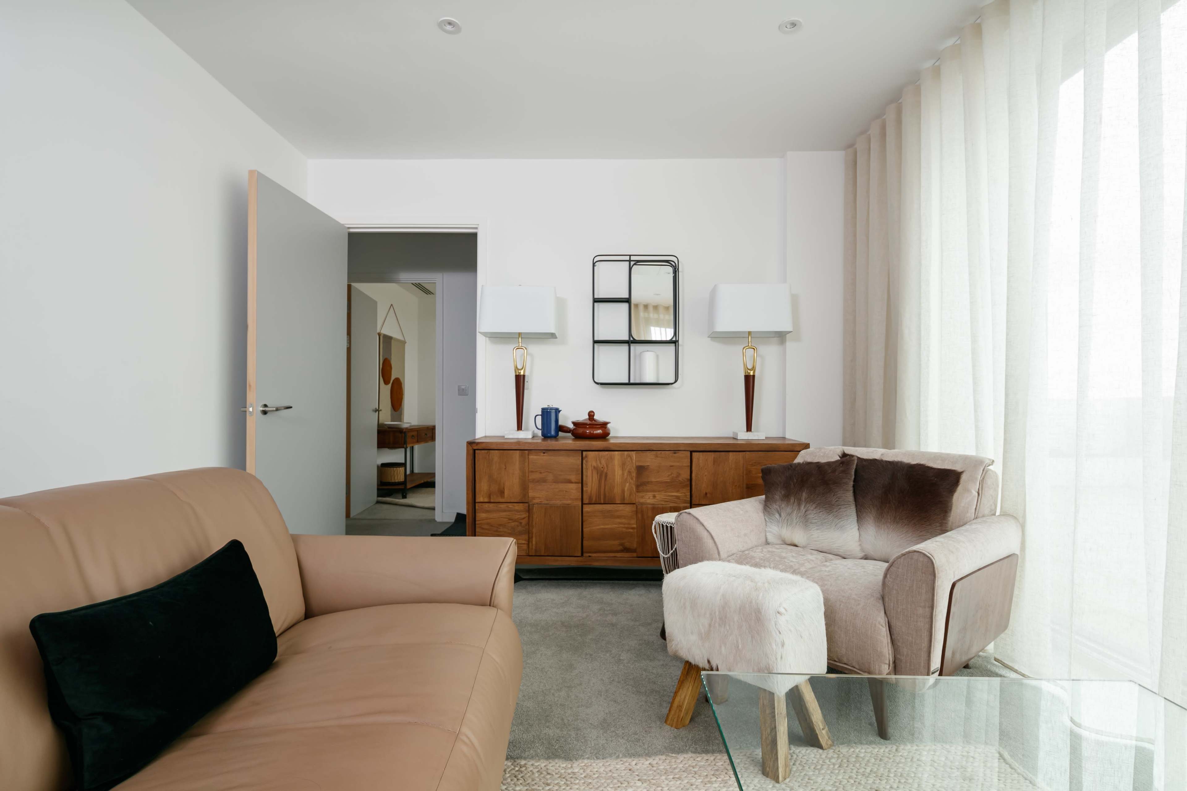 A modern living room with a tan leather sofa, a wooden sideboard, and large windows covered with sheer curtains.