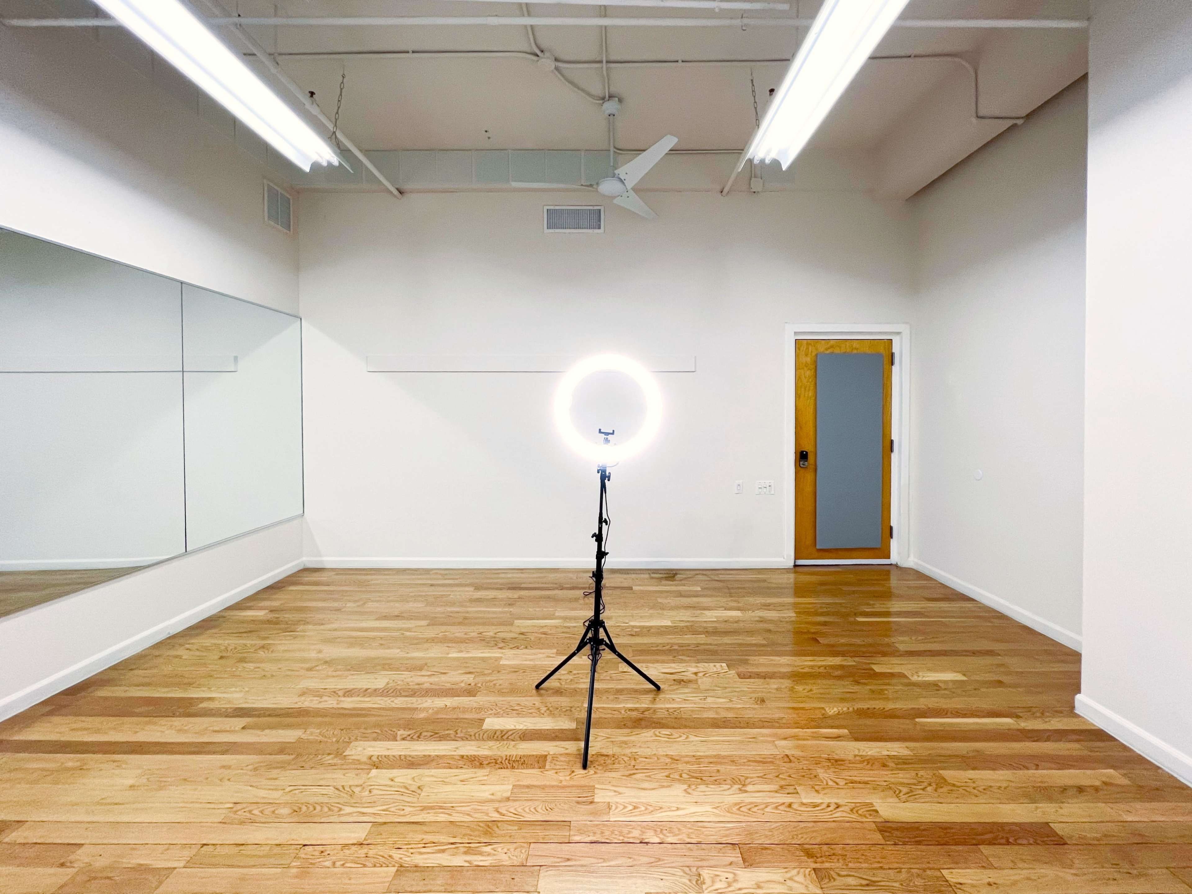 The image shows a well-lit, empty room with wooden floors, a freestanding ring light on a tripod, and a mirror along one wall.