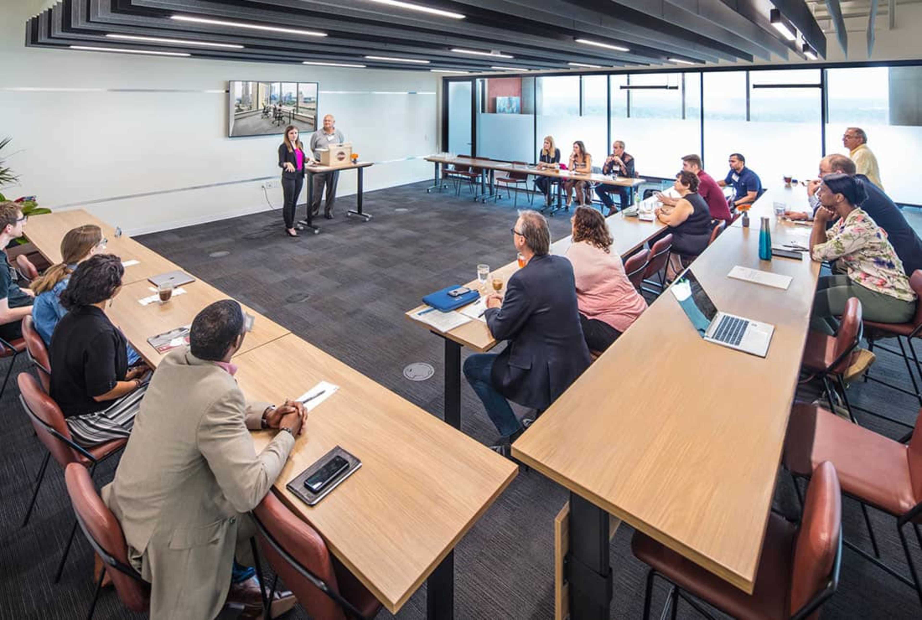 A group of people are gathered in a modern conference room seated around a long table, listening to a presentation being delivered at the front.