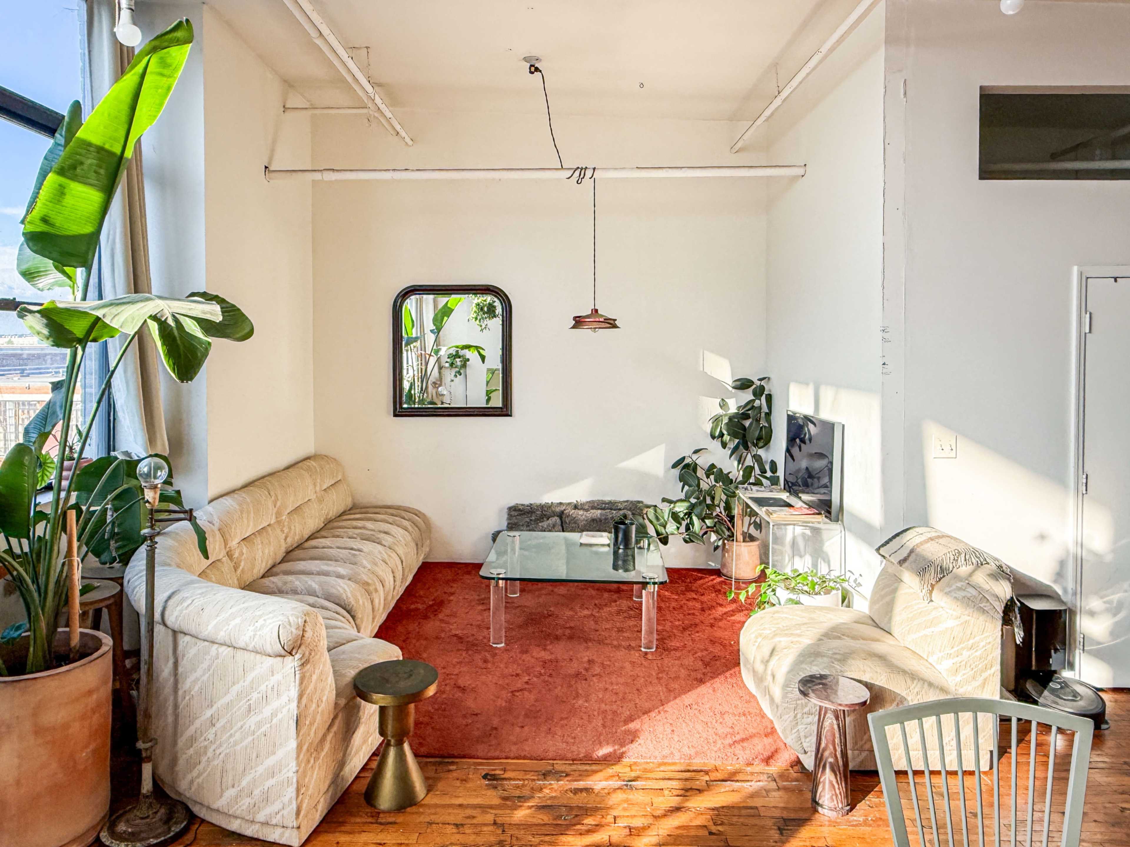 The image shows a living room with a large glass coffee table, a tan sectional sofa, and plants arranged around a red area rug.