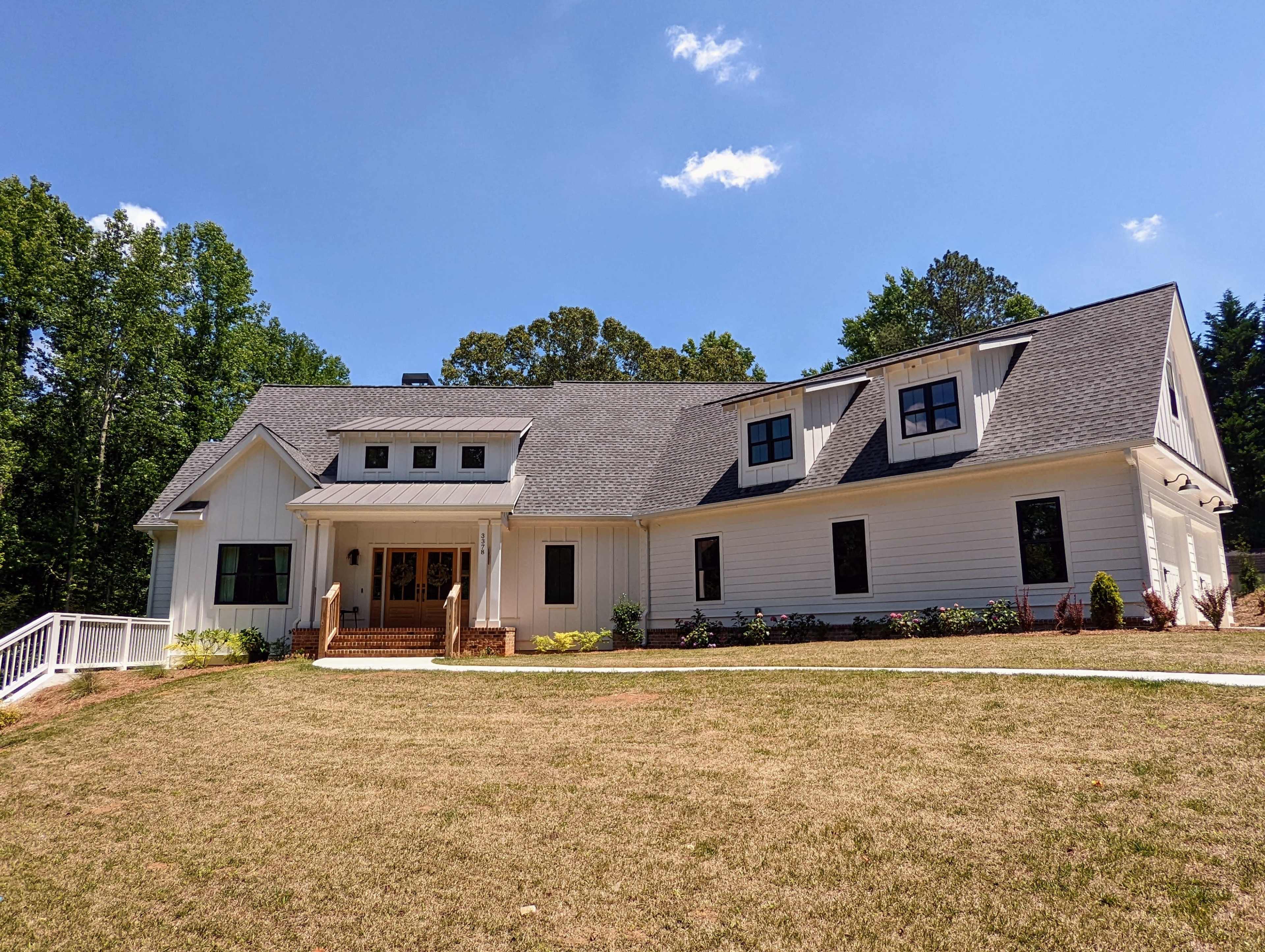 A large, modern white house with a gabled roof and multiple windows is situated on a grassy lot surrounded by trees.