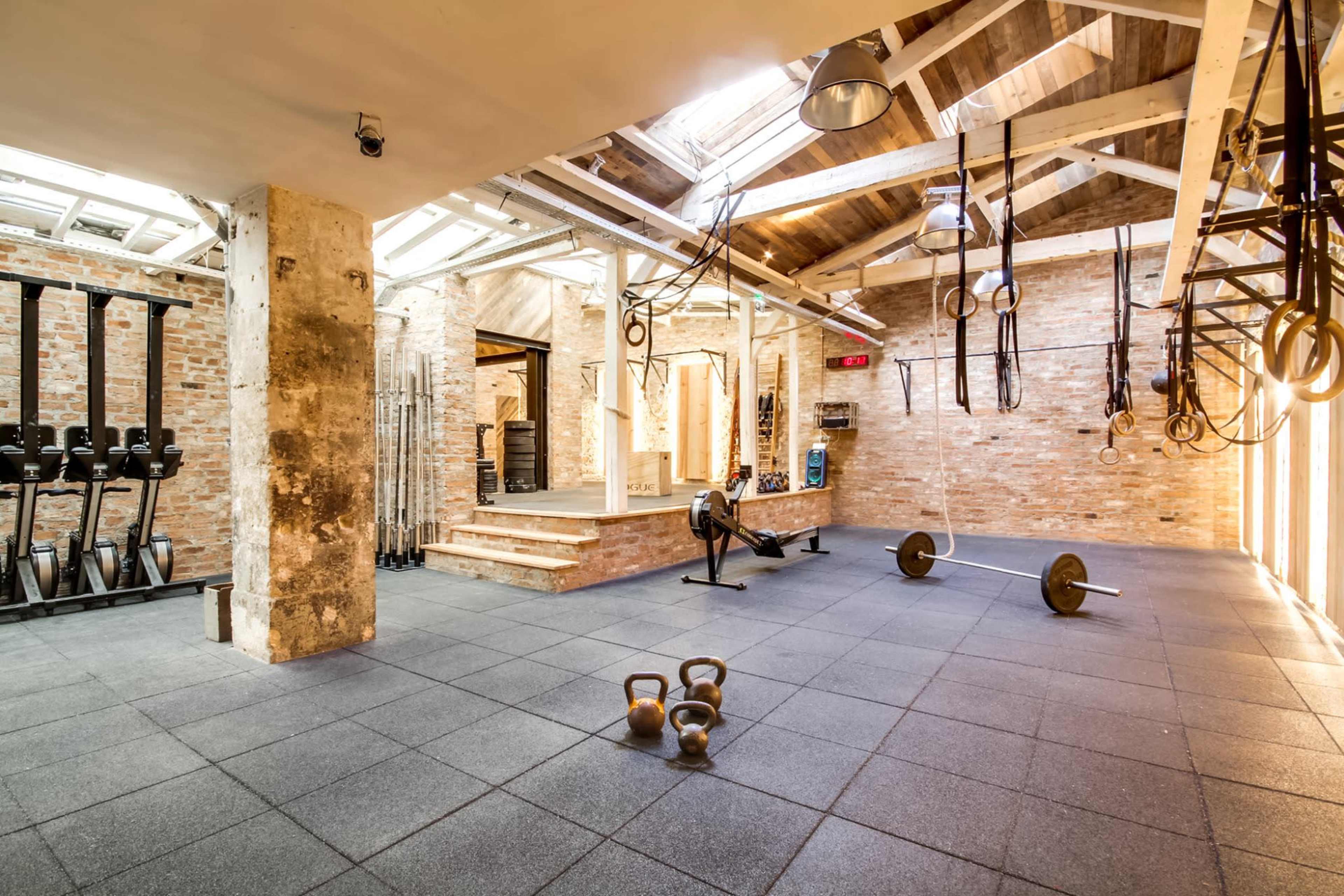 A spacious, industrial-style gym with exposed brick walls, various weightlifting equipment, and fitness tools arranged on a rubber floor.