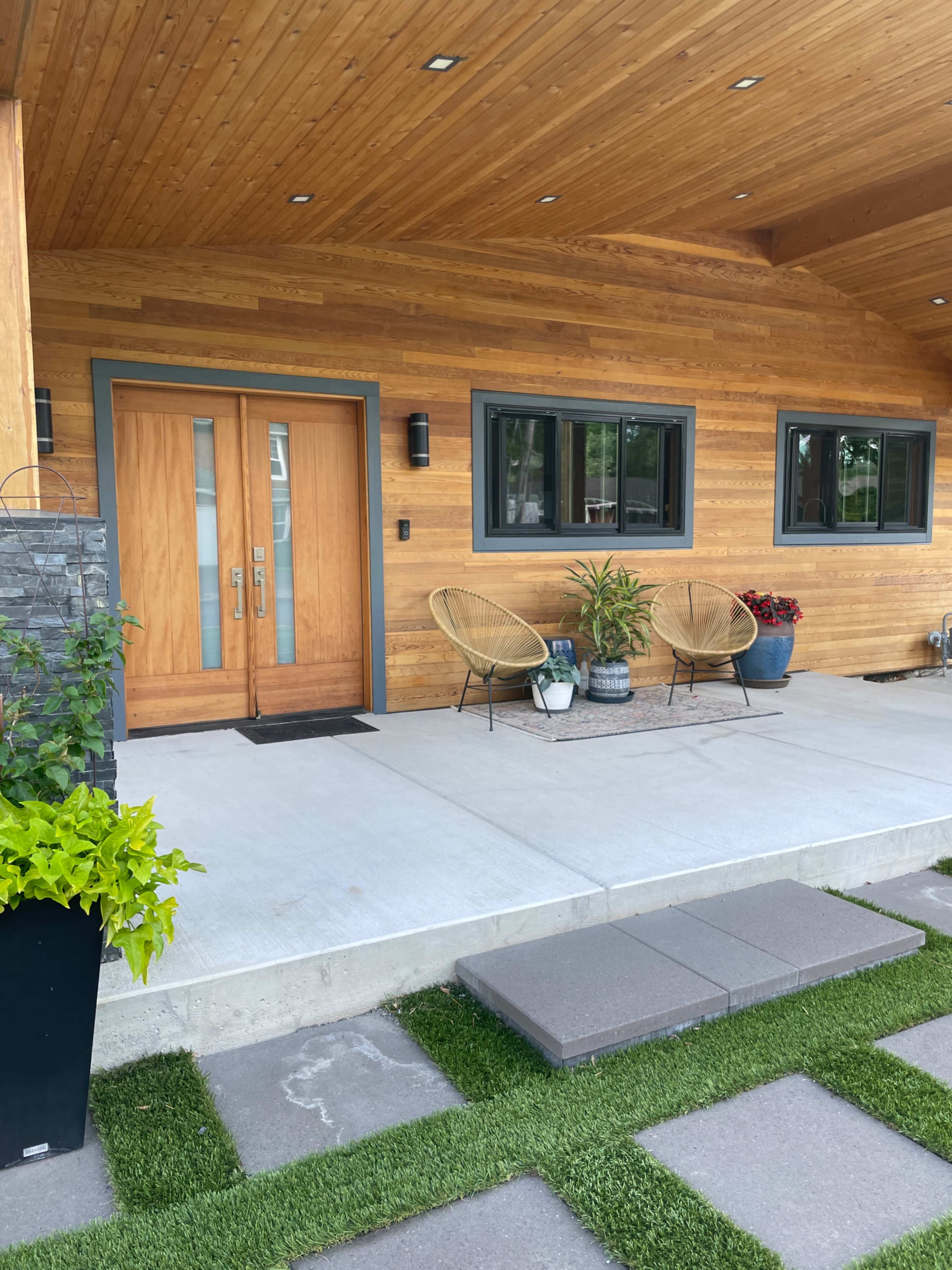 The entrance of a modern home with wooden siding, a concrete porch, and two wicker chairs beside a potted plant.