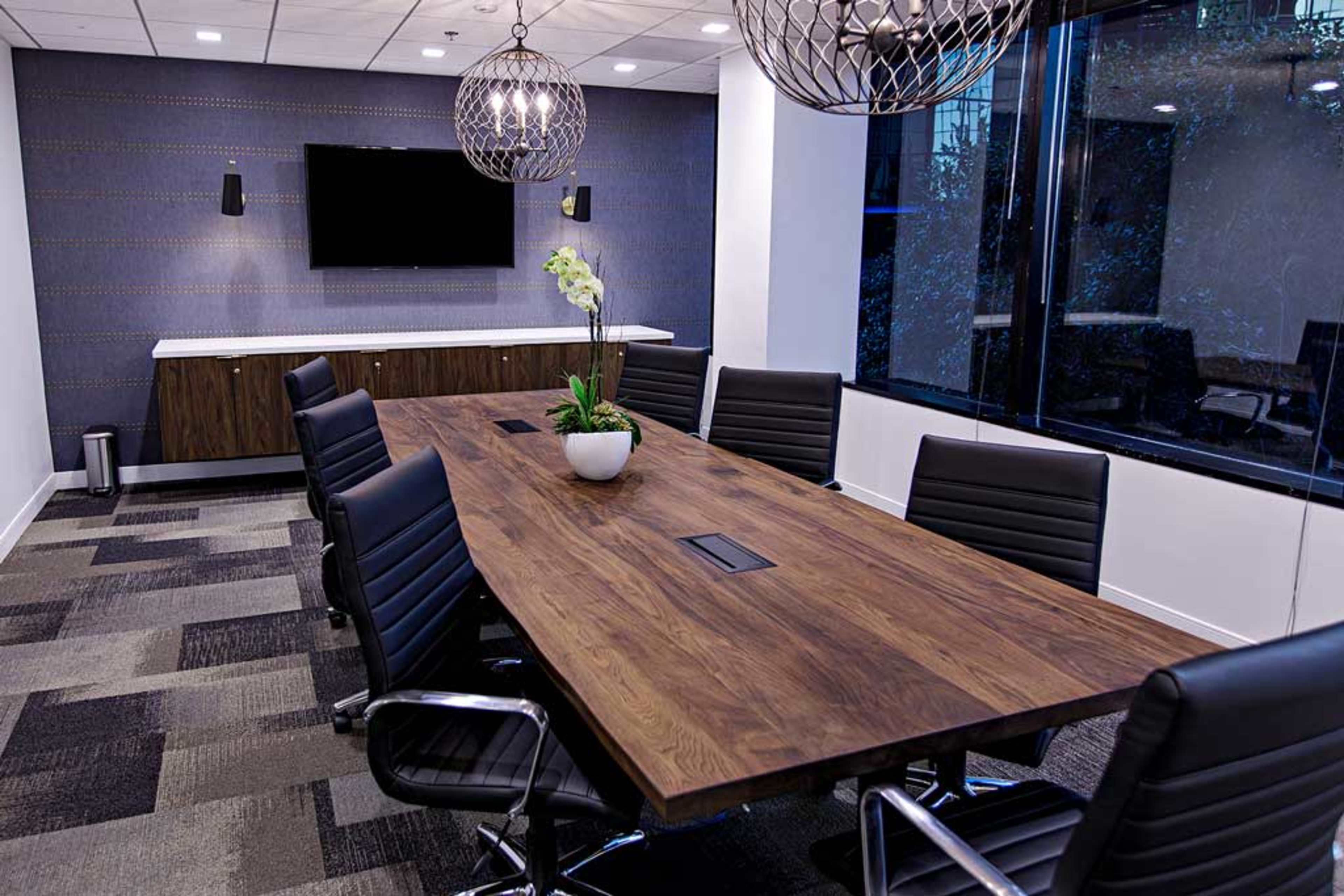 A large wooden conference table is surrounded by black leather chairs in a modern meeting room with a wall-mounted screen and large windows.