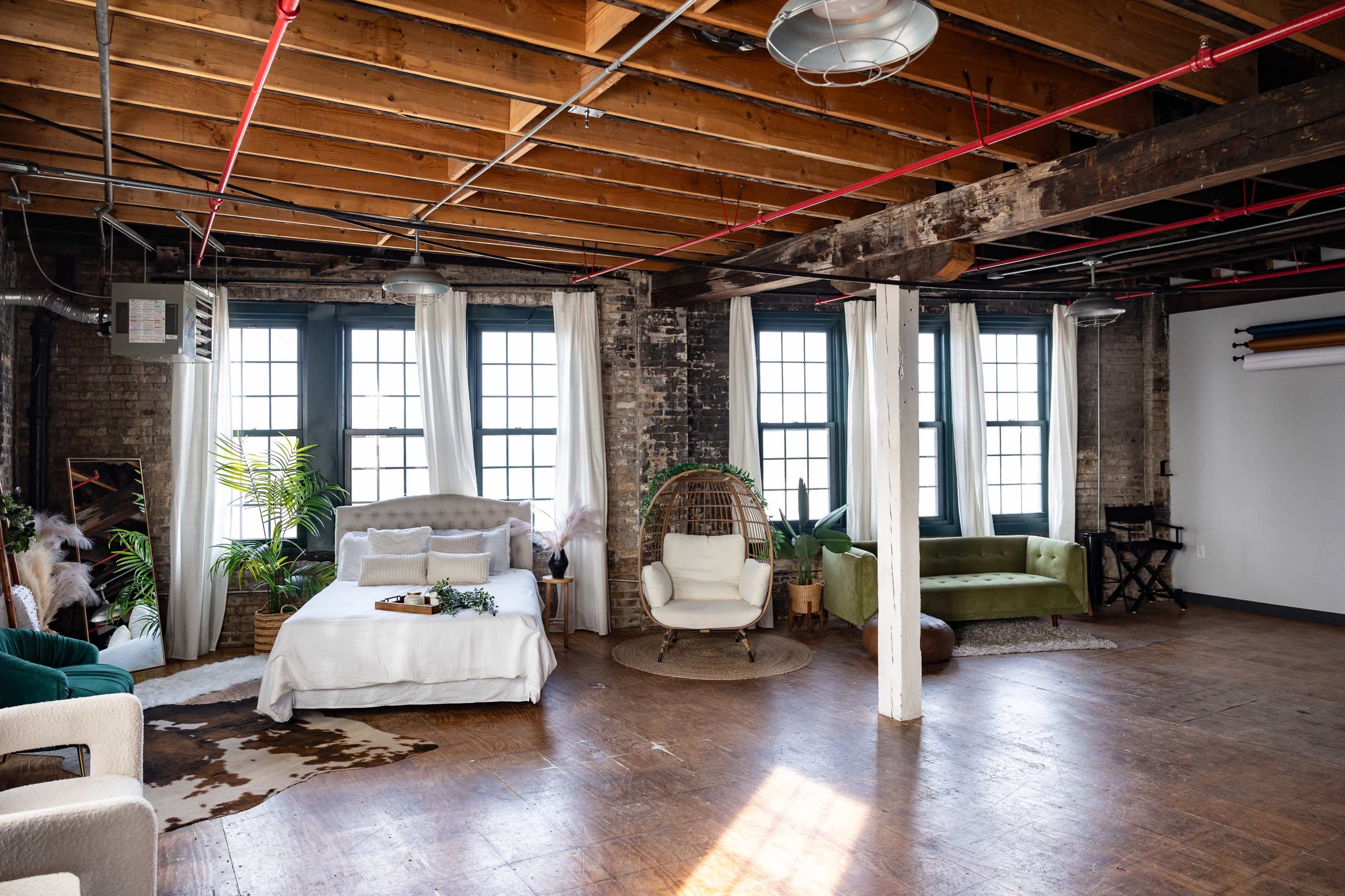 The image depicts an industrial-style studio apartment featuring a bed, a green couch, a white chair, and large windows with natural light streaming in.