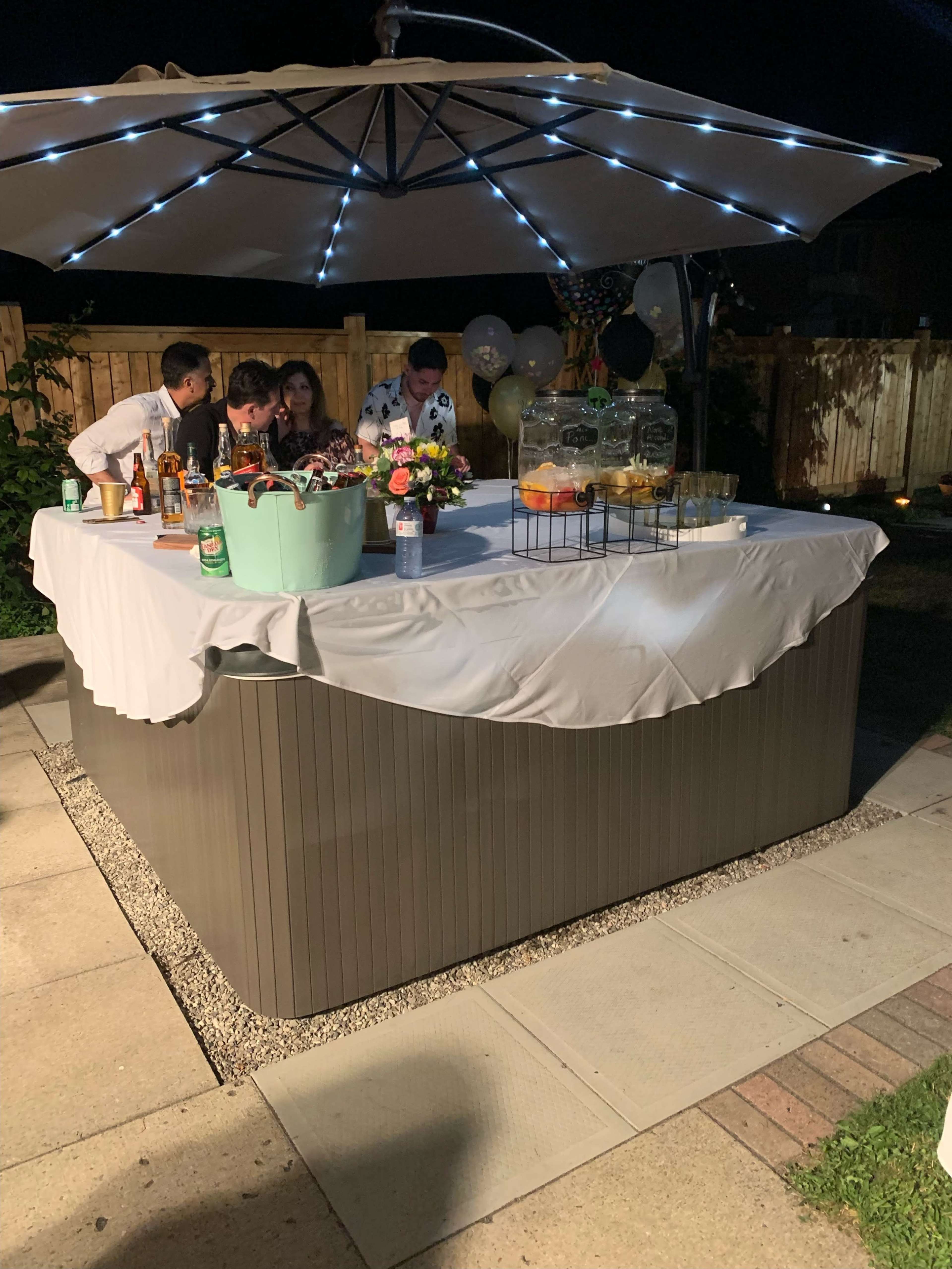 A group of people gather around a bar setup with drinks and decorations under an umbrella in a backyard setting at night.