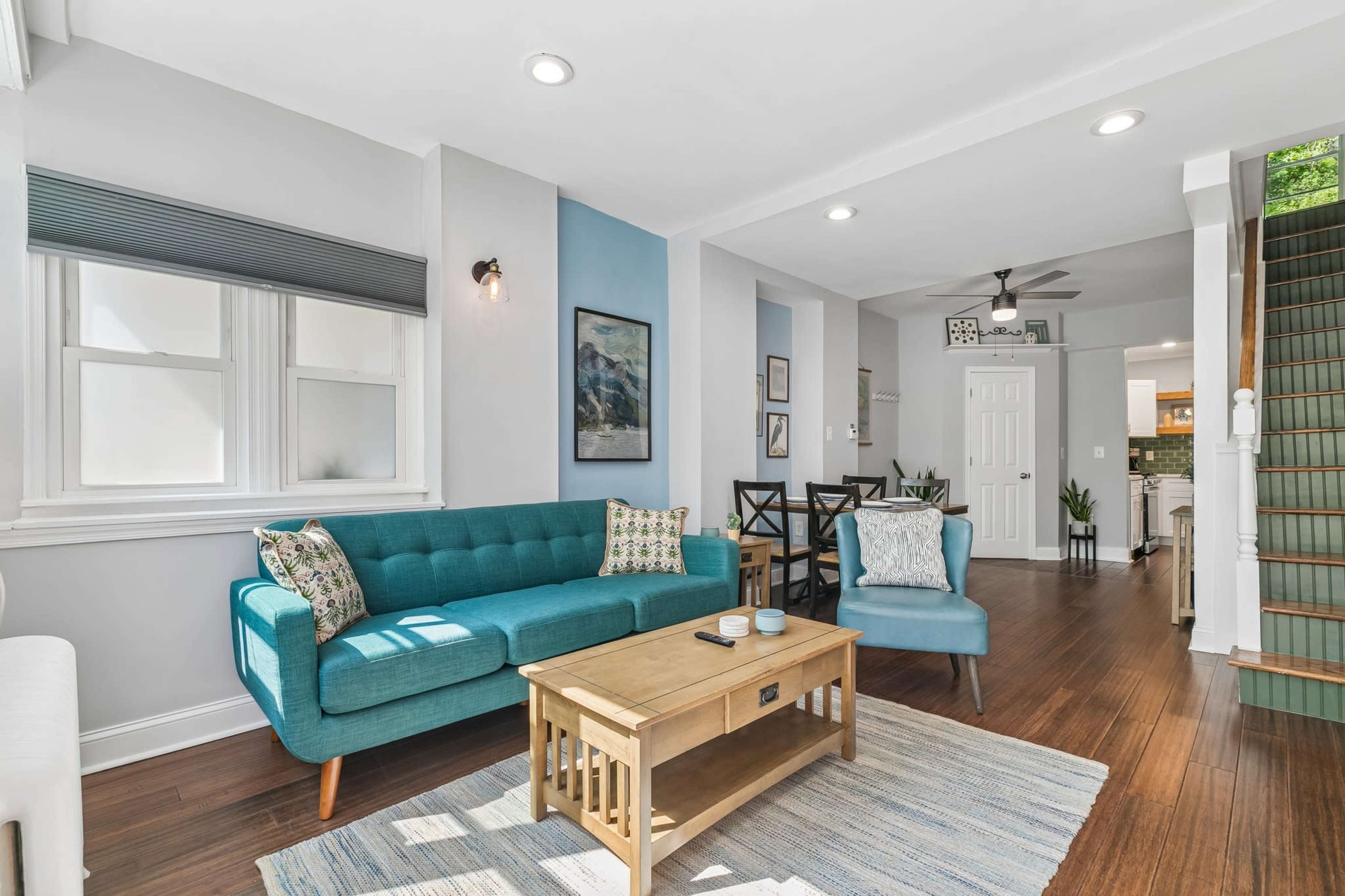 The image shows a bright living room with a teal sofa, a wooden coffee table, and a staircase leading to another level, with a dining area visible in the background.