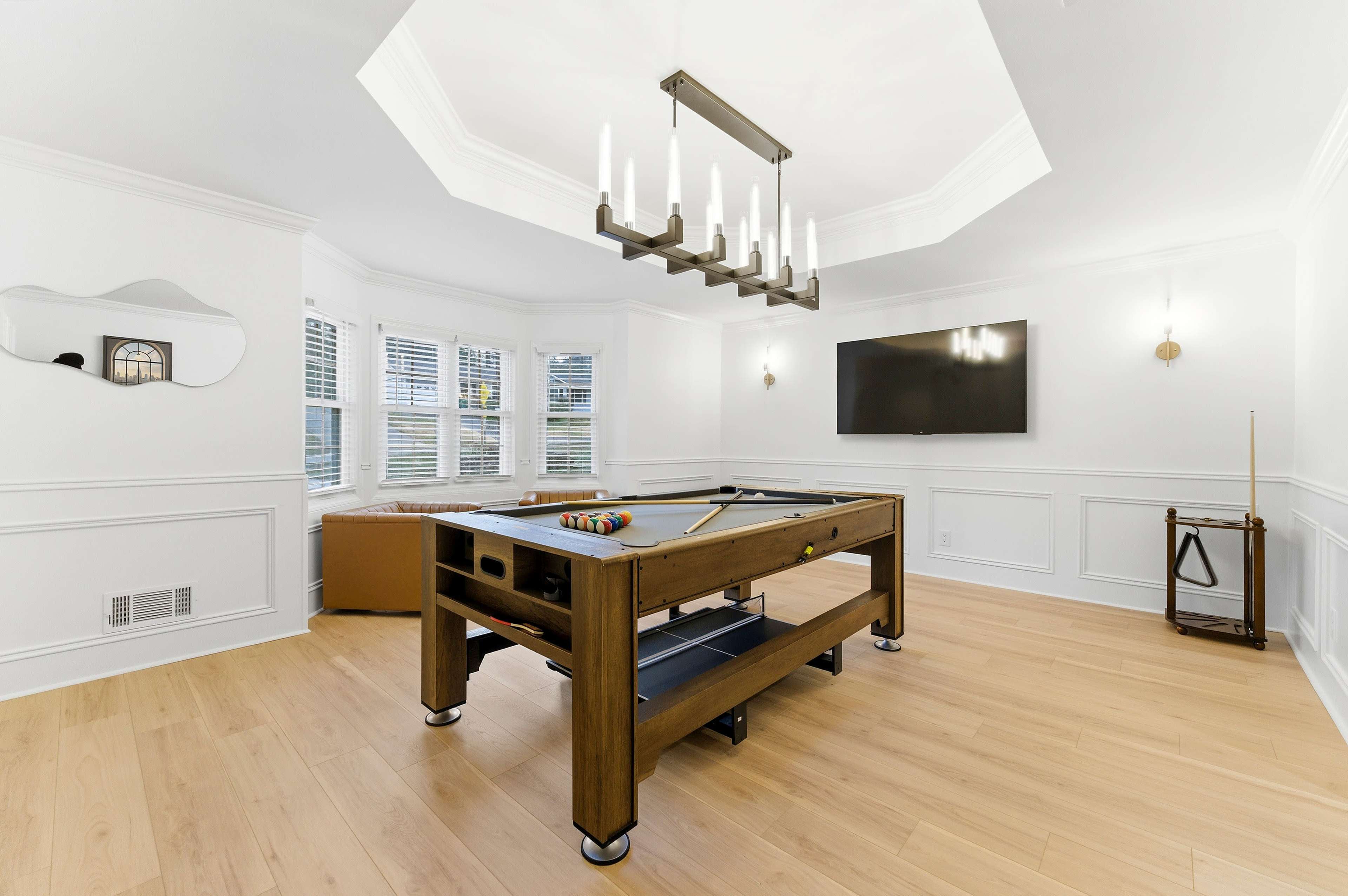 The image shows a bright game room featuring a pool table, a modern light fixture, a wall-mounted TV, and large windows with white trim.