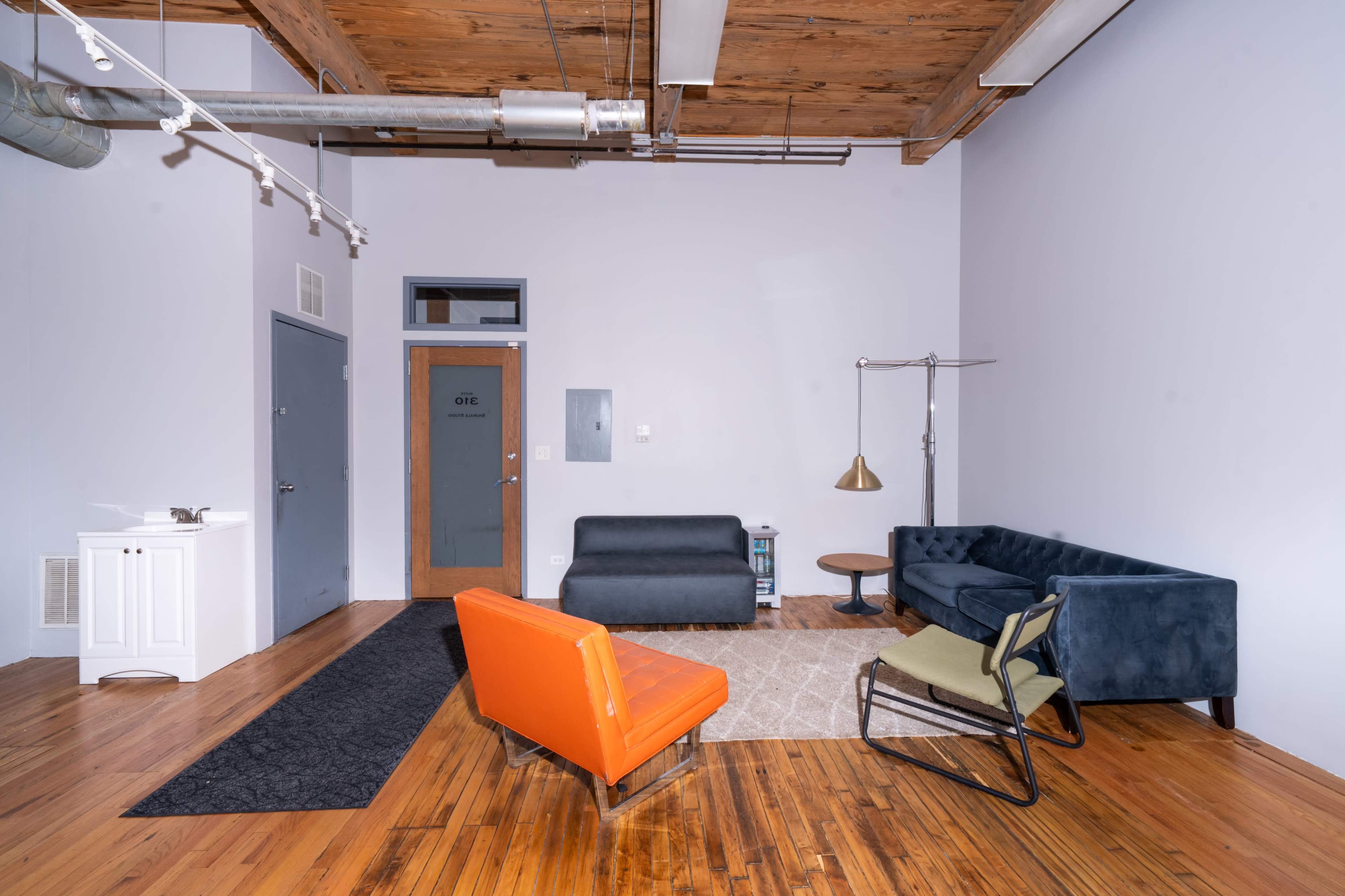 The image shows a spacious room with wooden floors, featuring a blue sofa, an orange chair, a green chair, a lamp, and a door labeled "OFFICE."