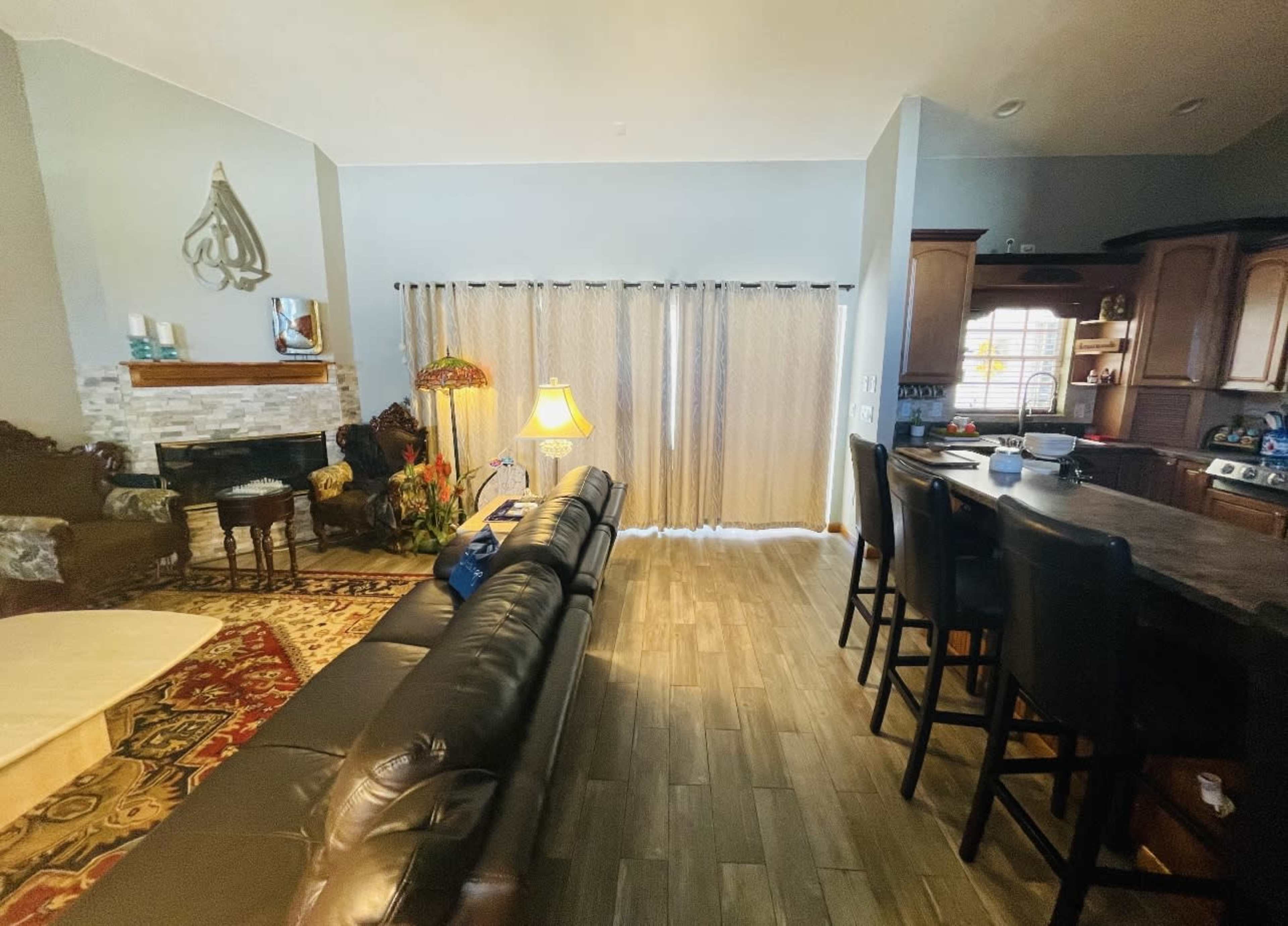 A living room with a sofa, a coffee table, and a kitchen area visible in the background, featuring a bar with stools and a fireplace.