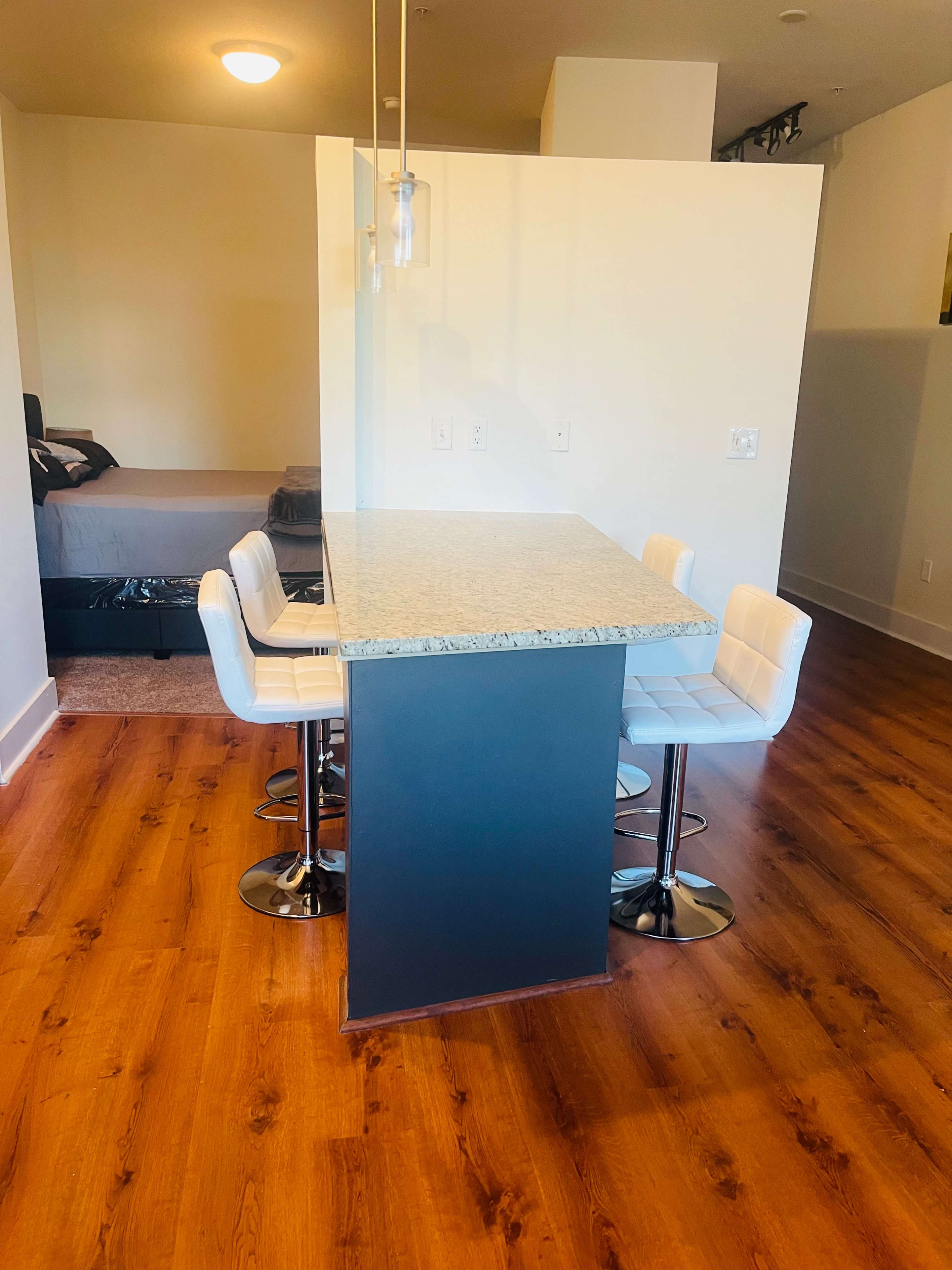 A kitchen island with granite countertop and four white bar stools is positioned in front of a blurred living area in a modern apartment.