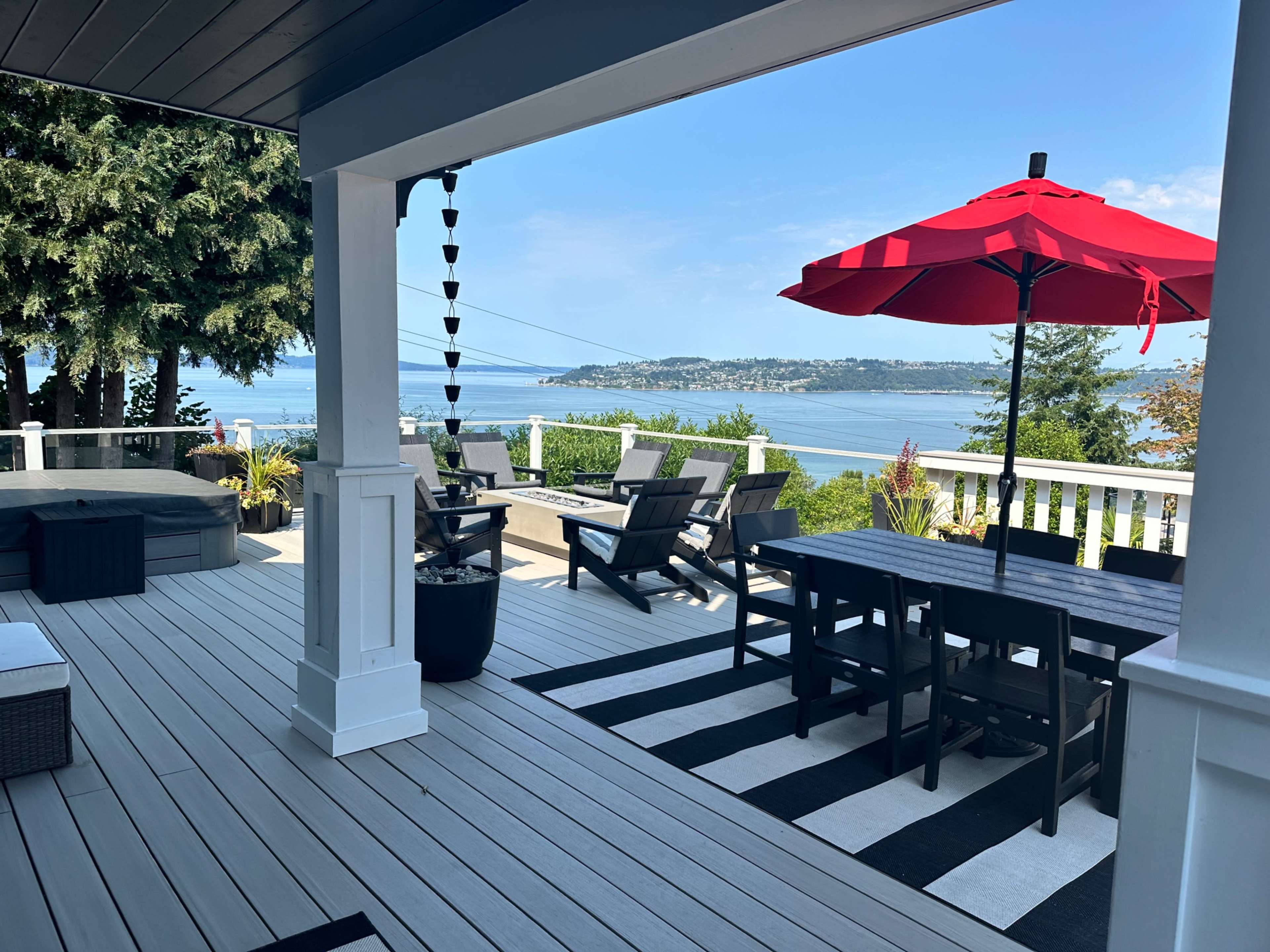The image shows a patio overlooking a body of water, featuring a dining table with chairs, lounge chairs, and a large red umbrella.