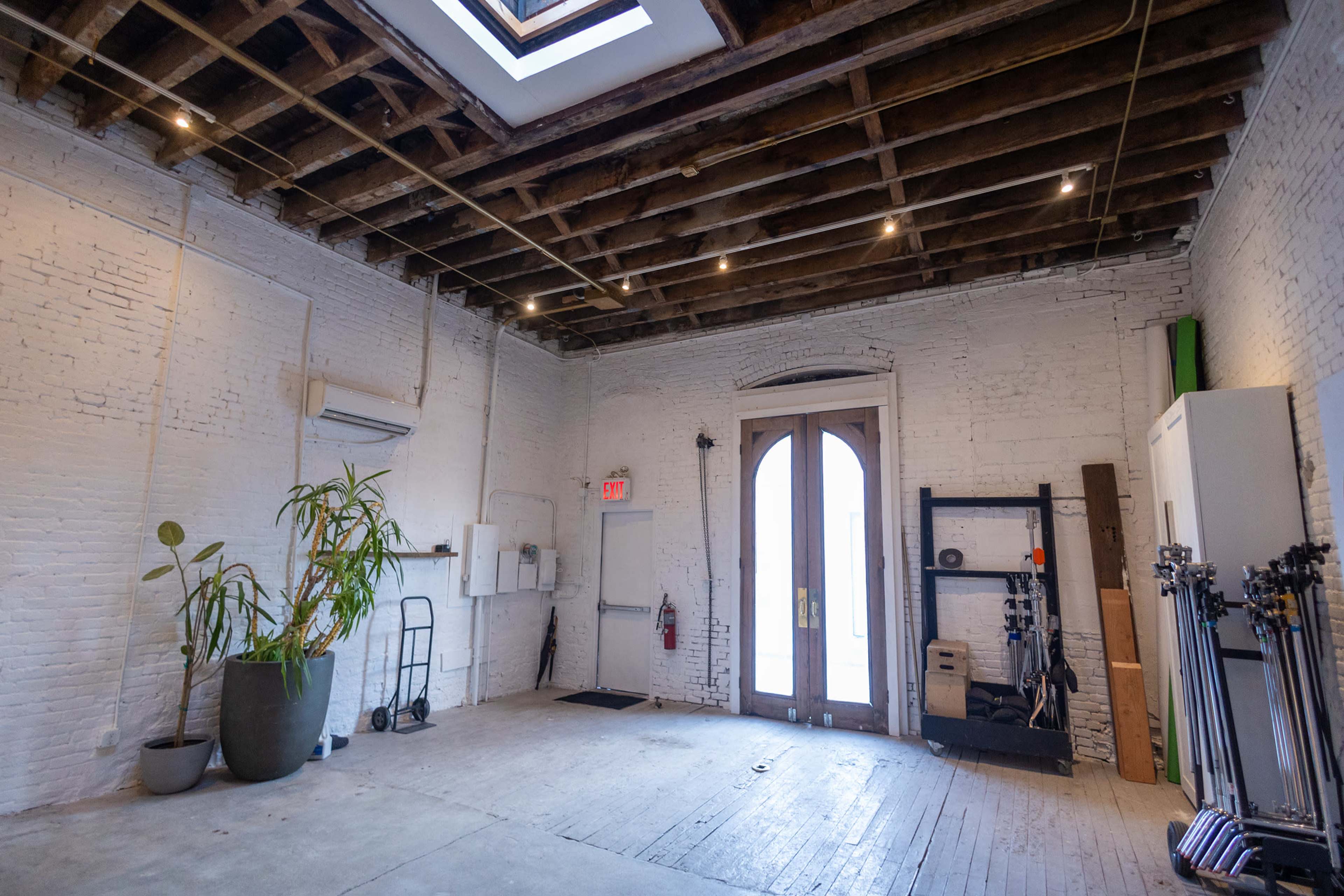 An empty room with exposed wooden beams, white brick walls, and large double doors leading outside, along with a few plants and equipment along the walls.