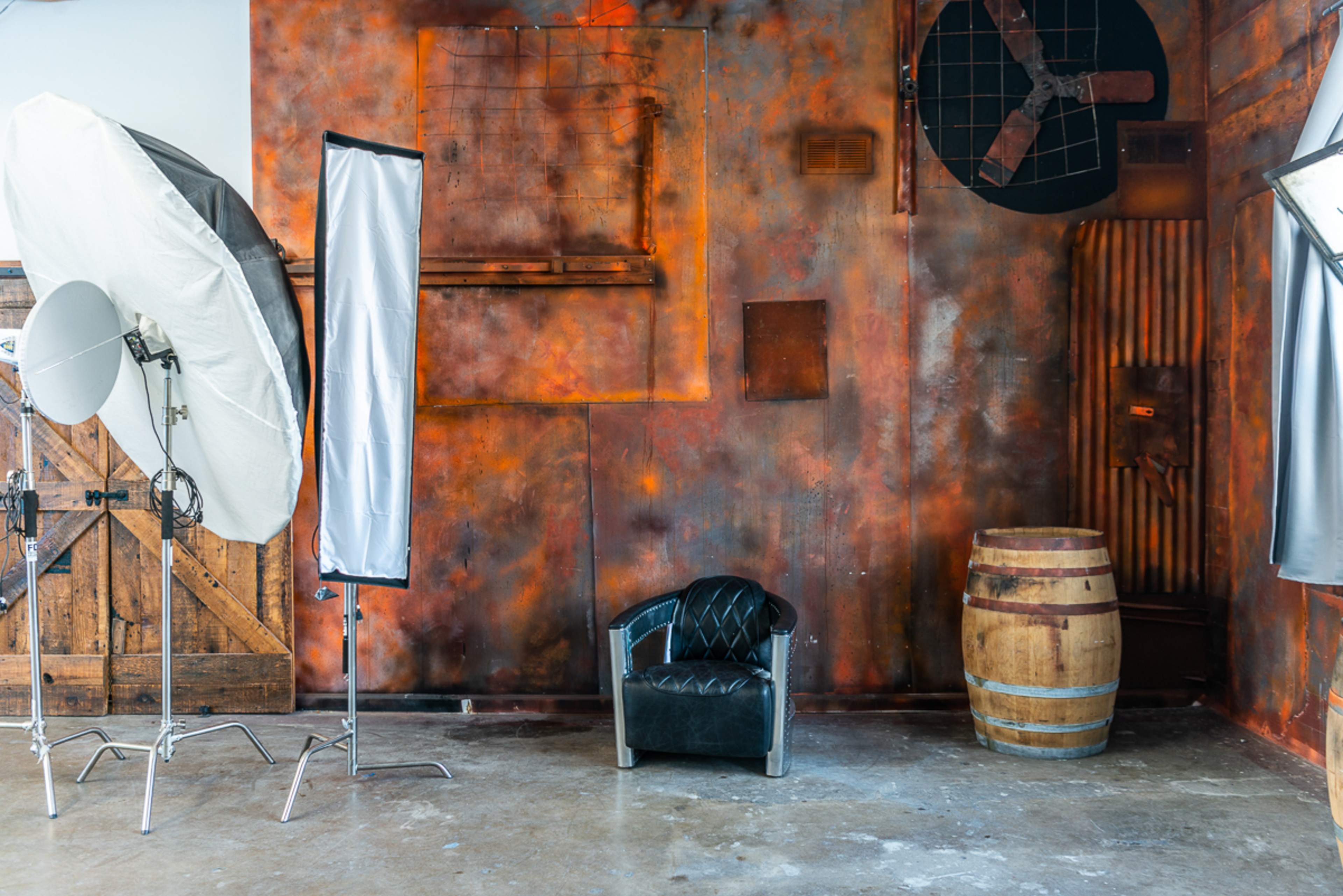 The scene features a photography studio with a textured, orange-toned wall, a black leather chair, a wooden barrel, and two light setups.