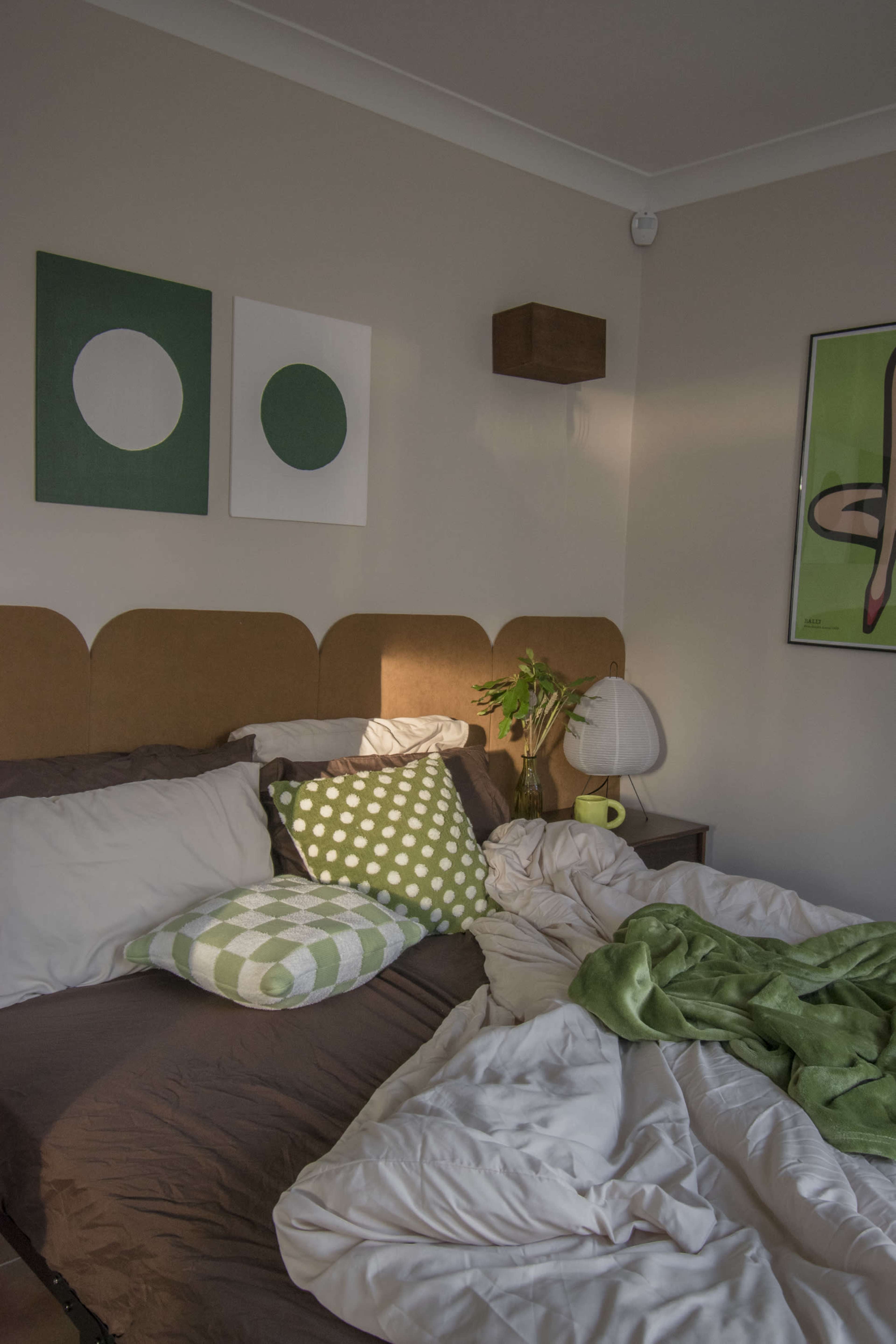 A neatly made bed with a green blanket and patterned pillows is positioned against a wall adorned with abstract art.