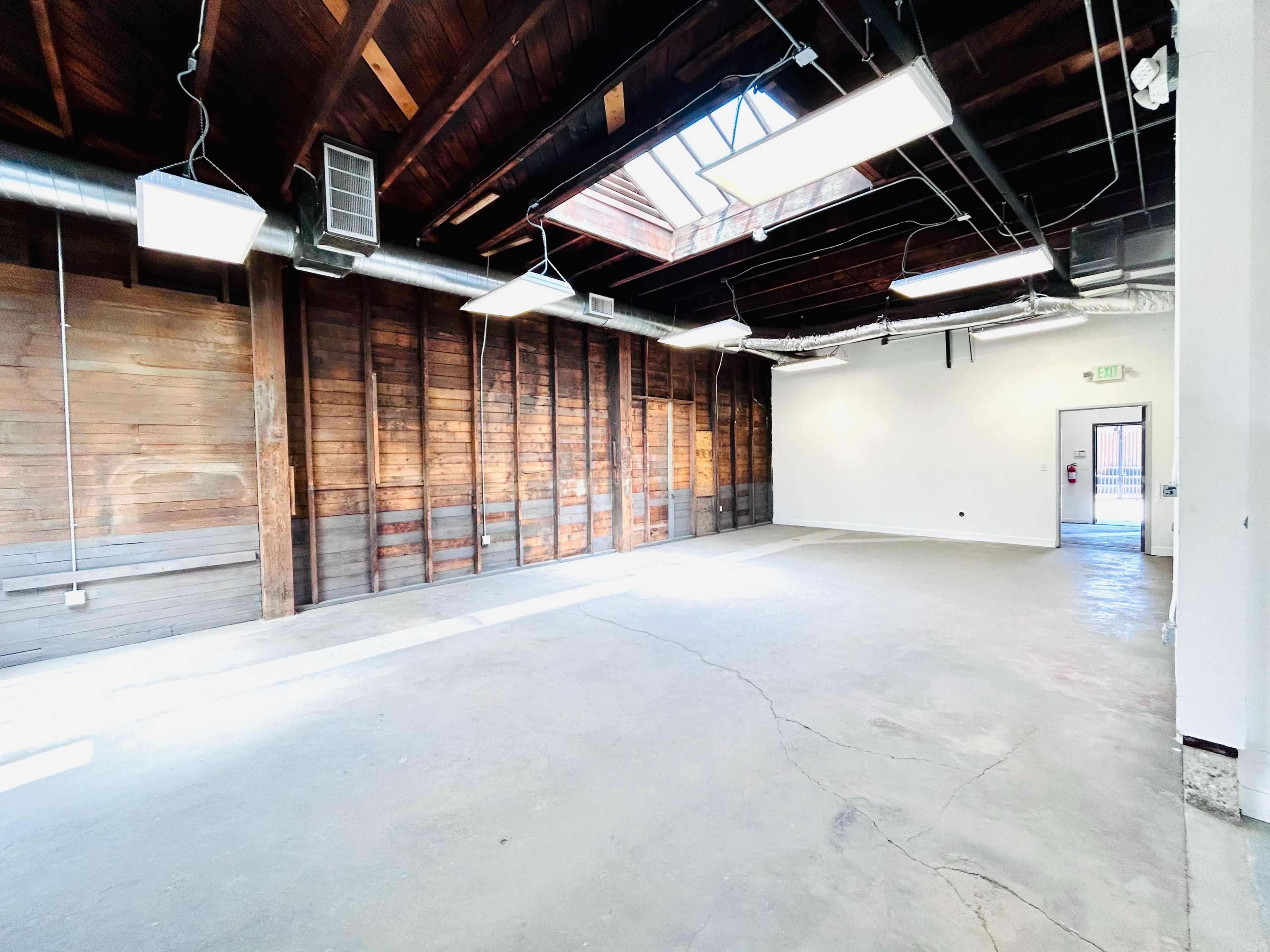 The image shows a spacious, vacant room with wooden walls, large skylights, and concrete flooring.