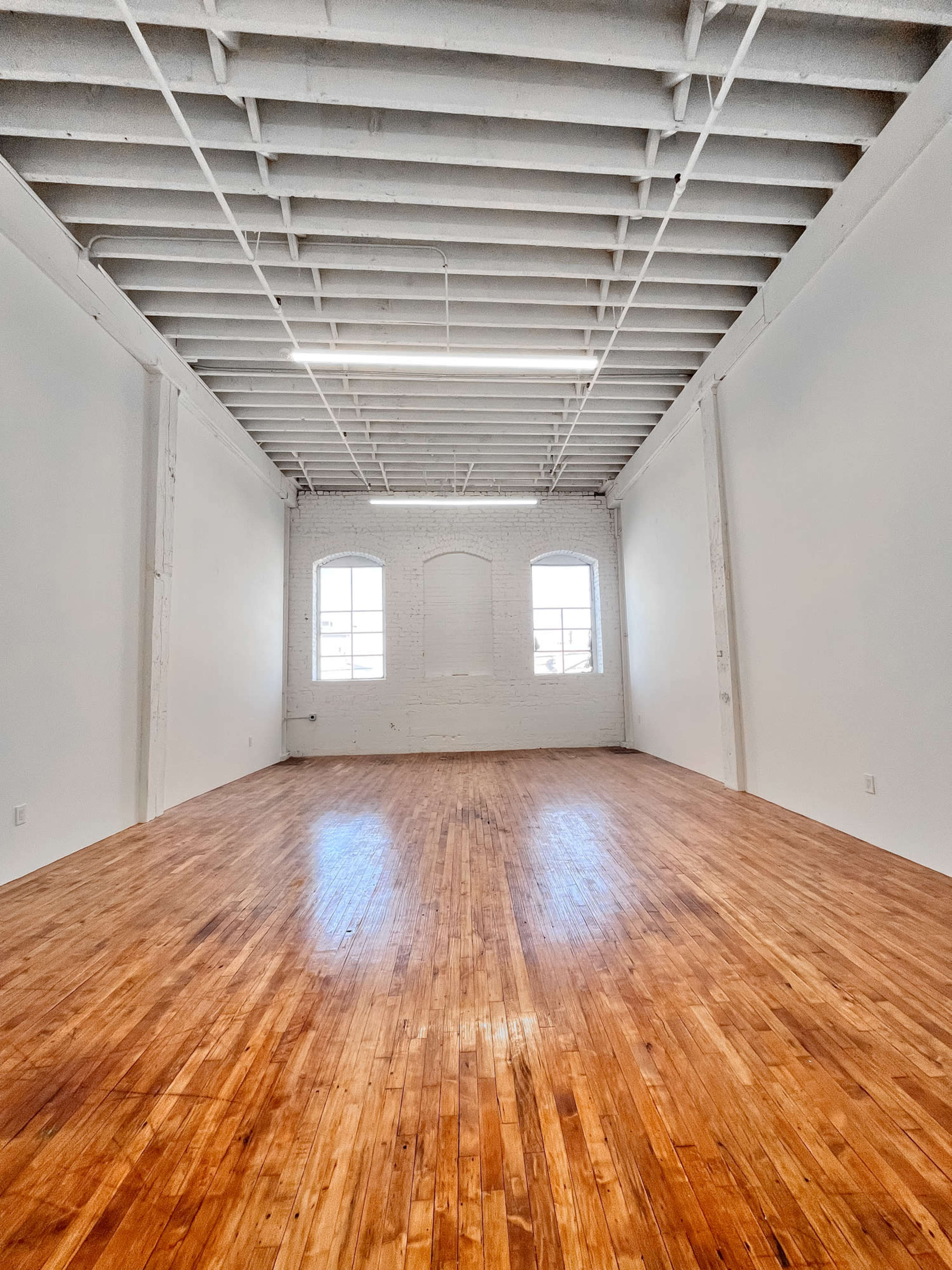 A spacious, empty room with hardwood floors and white walls, featuring large windows at one end.