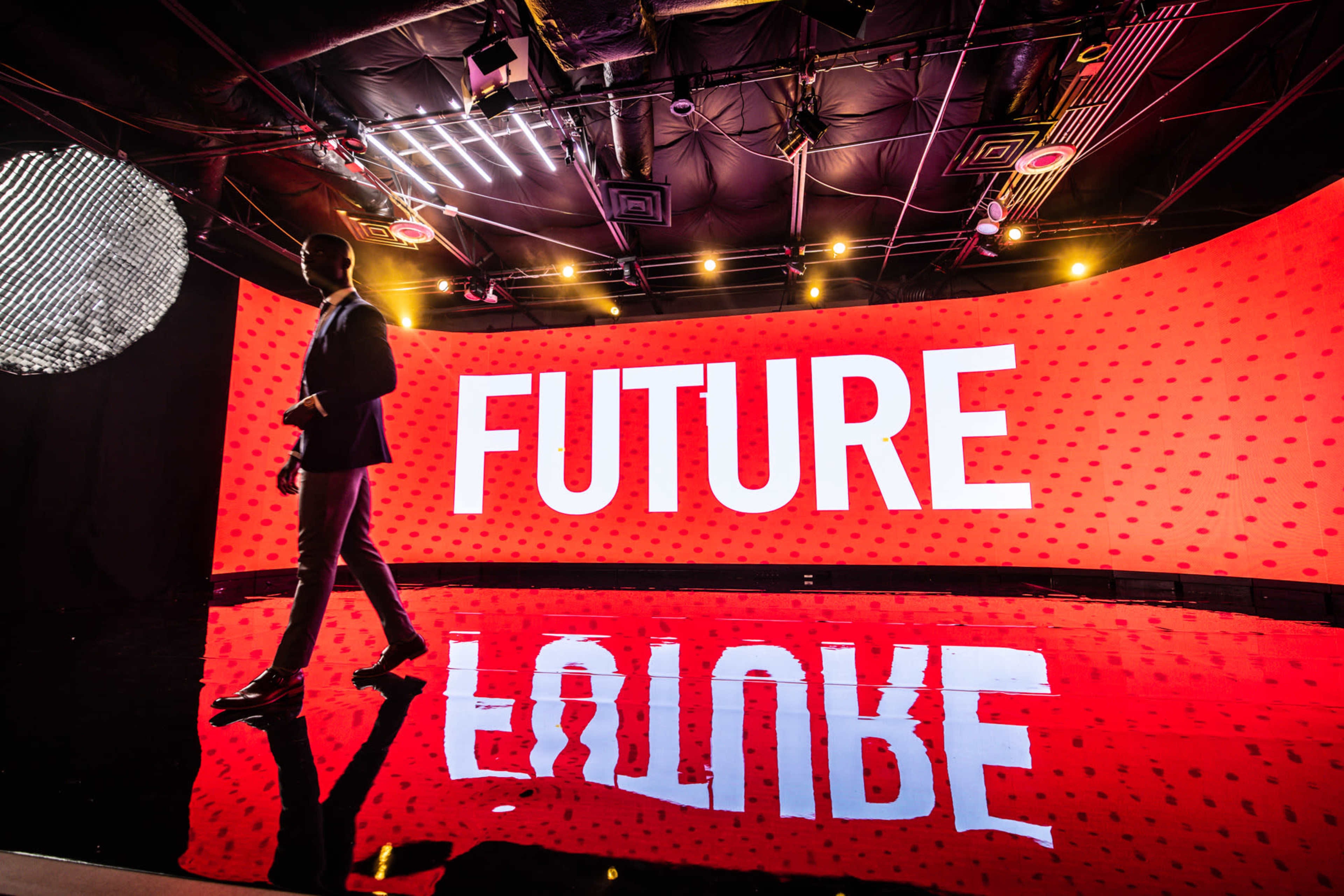 A figure walks on a reflective stage with a large illuminated backdrop displaying the word "FUTURE" against a red background.