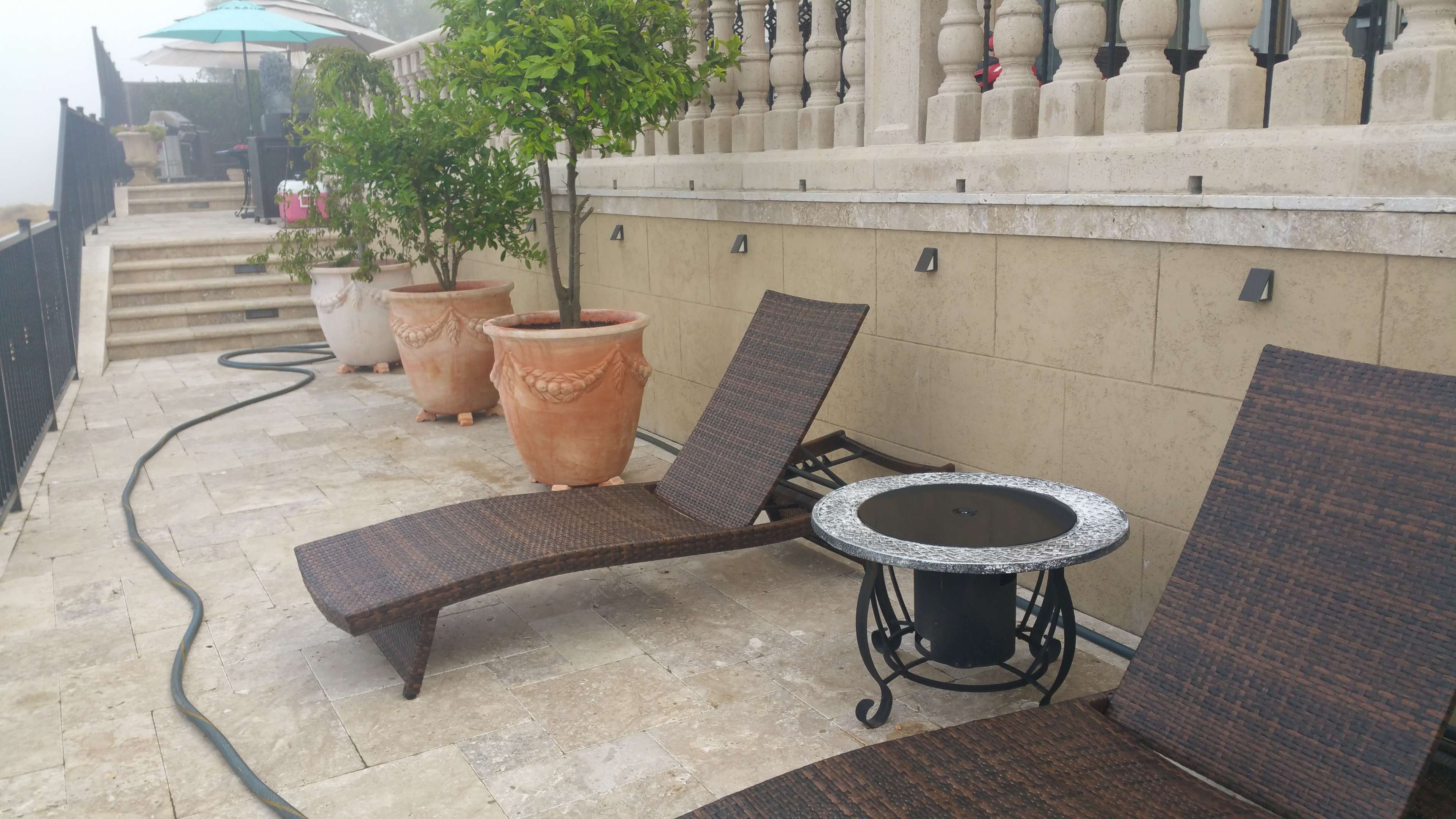 A stone patio with two lounge chairs, a small round table, and several large potted plants near a staircase.