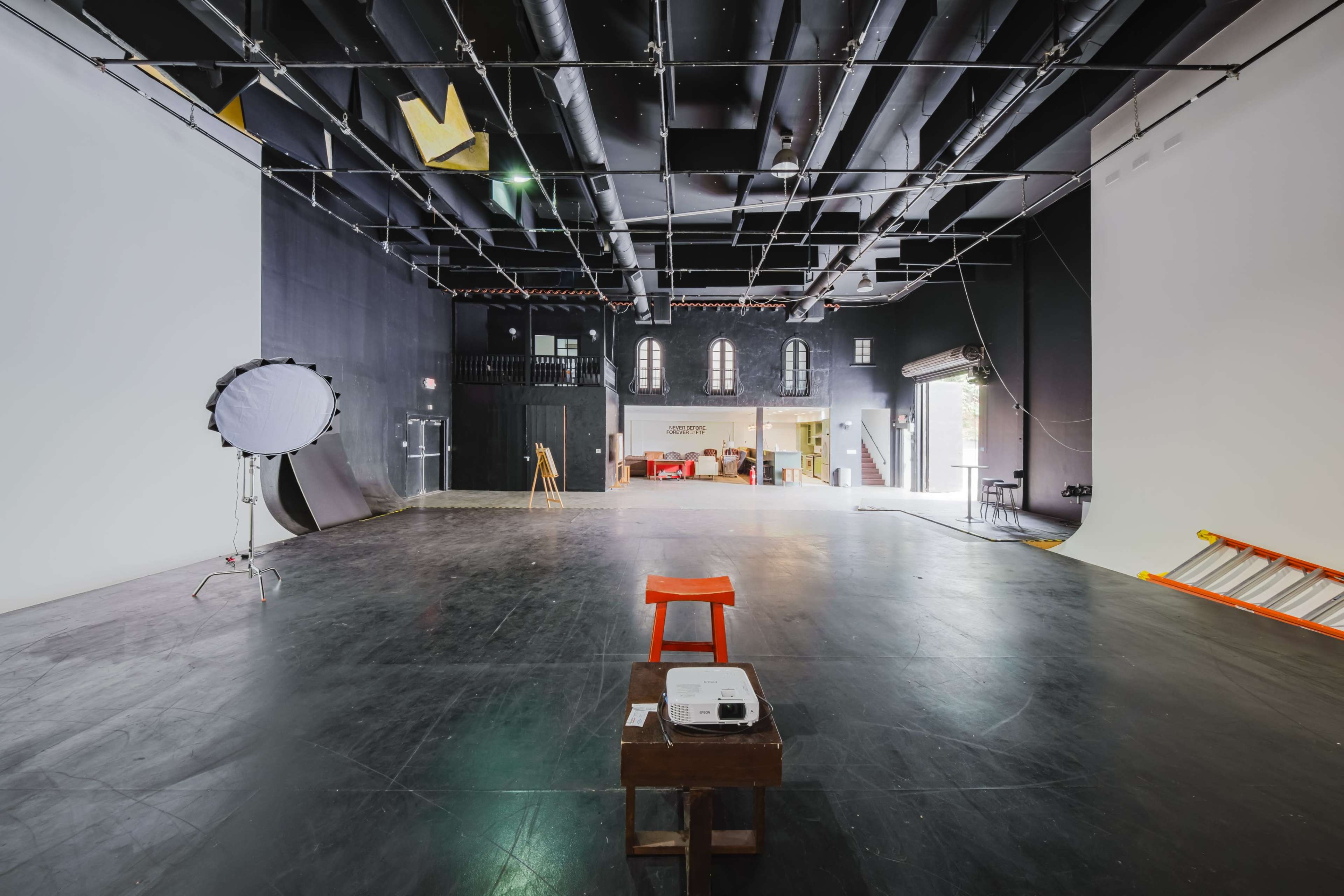 The image shows a spacious film studio with a black floor, white and black backdrops, scattered lighting equipment, and a small seating area.