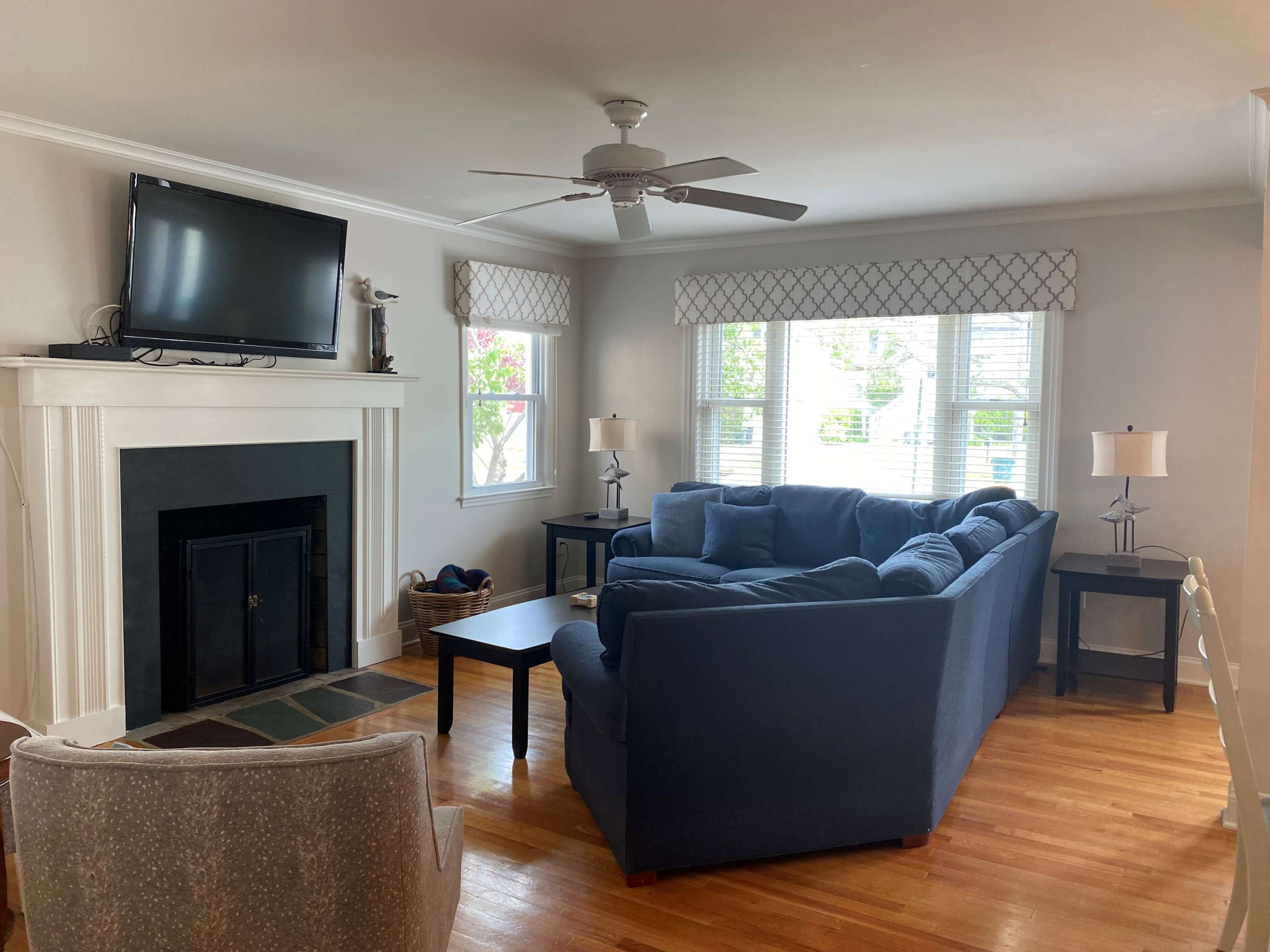 A living room features a blue sectional sofa facing a black fireplace and a television mounted above it, with windows dressed in patterned shades and two side tables with lamps.
