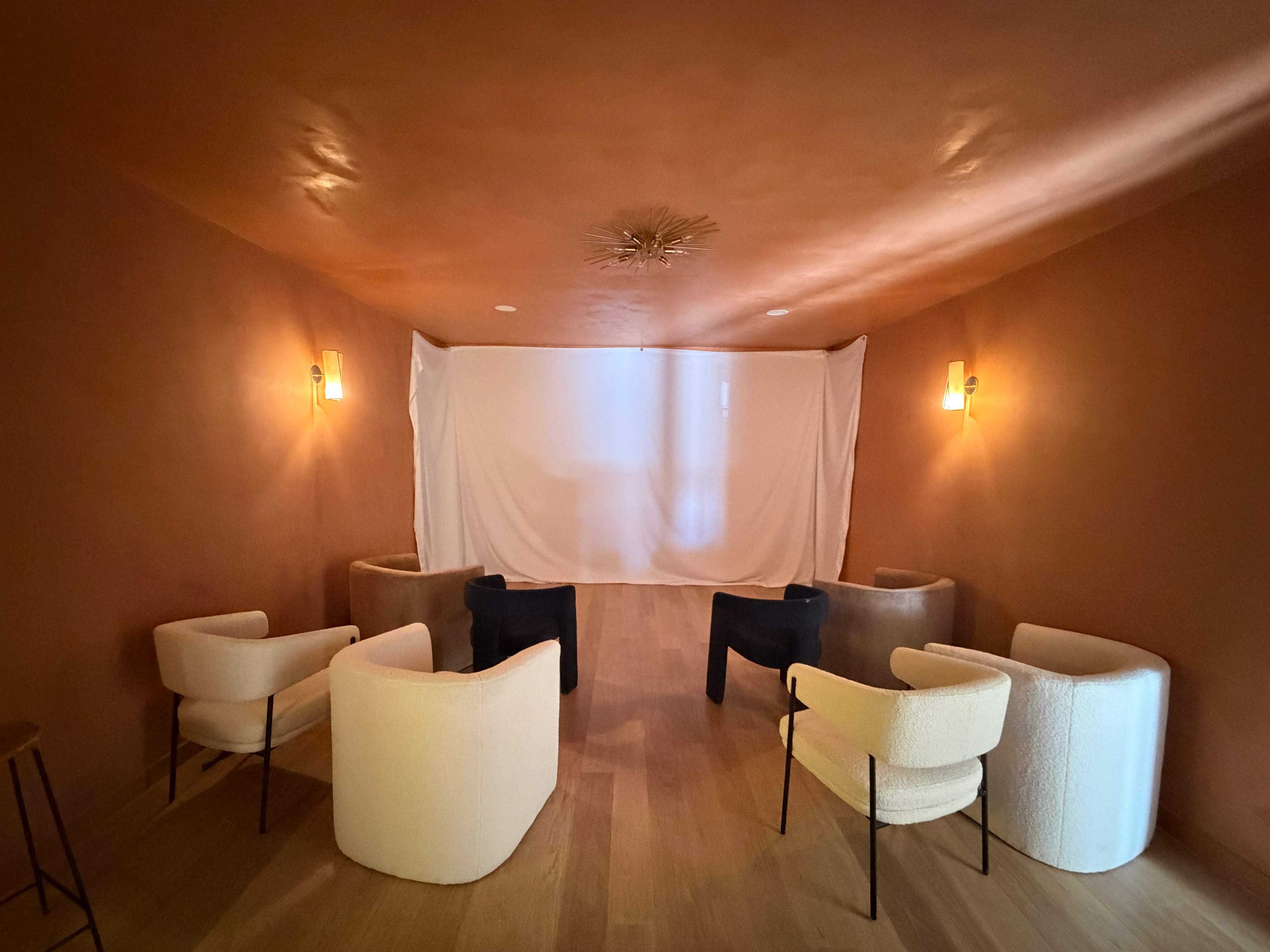 The room features a minimalist design with several seating chairs arranged around a white backdrop, illuminated by wall sconces on the orange-brown walls.