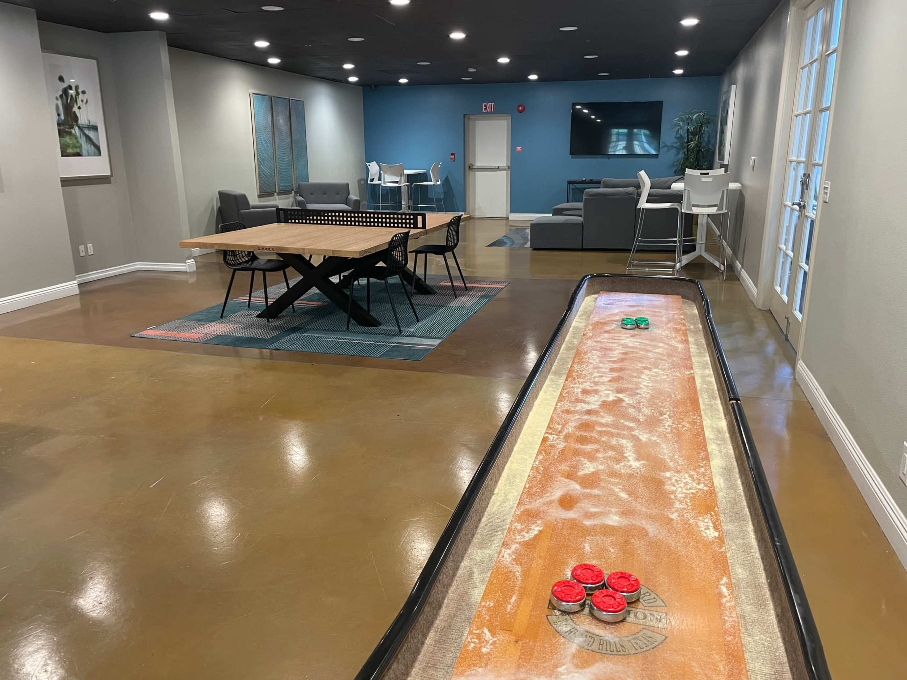 A recreational room featuring a shuffleboard table, a communal dining table, seating areas, and a large TV against the wall.