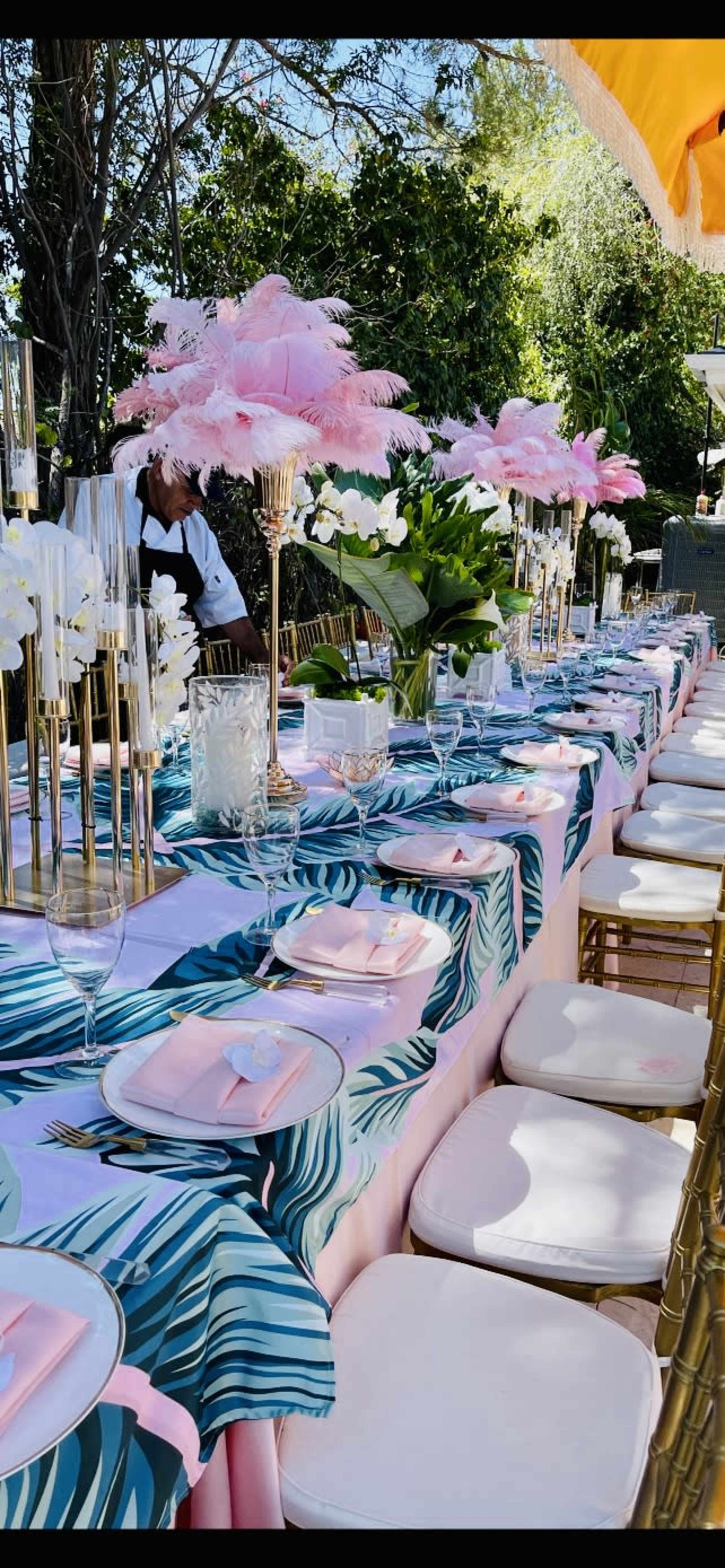A long dining table is decorated with elegant floral centerpieces, pink napkins, and a tropical-themed tablecloth, set in an outdoor garden setting.