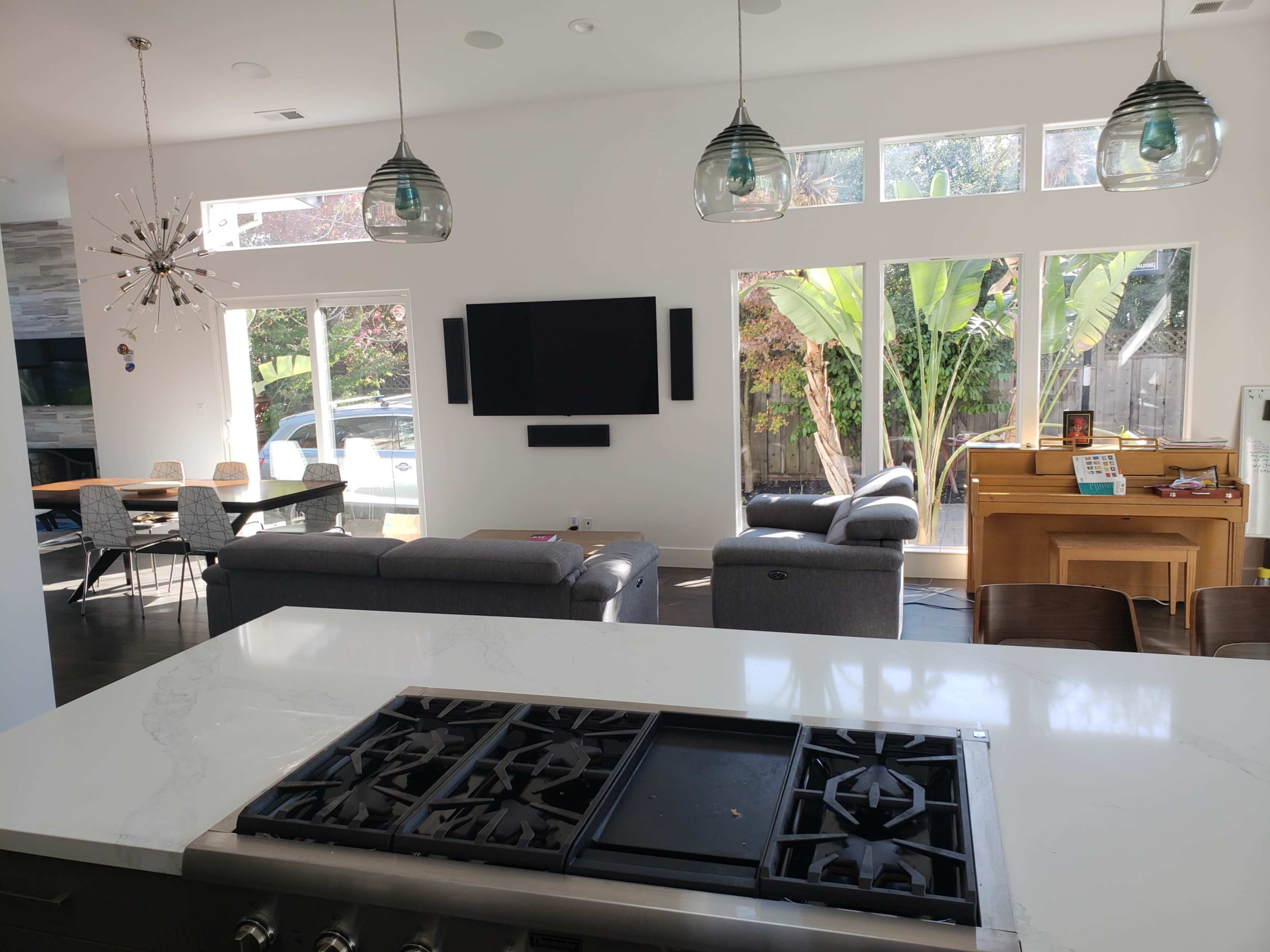A modern open-concept living space features a kitchen island with a gas range, a seating area with gray sofas, a wooden dining table, and large windows that overlook a patio with lush greenery.
