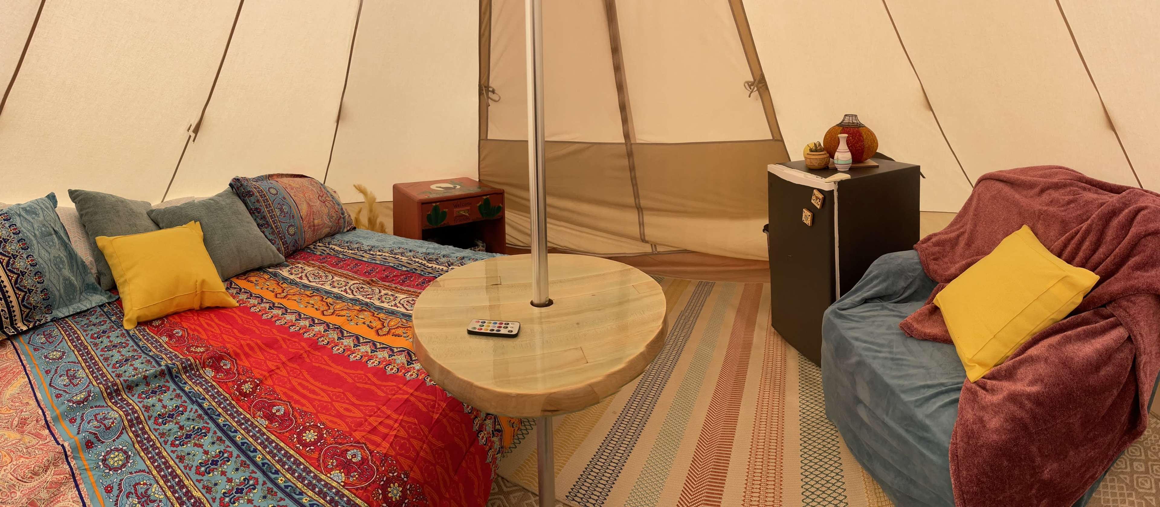 The interior of a tent features a colorful bedspread, a round wooden table with a remote, a small fridge, and comfortable seating areas.