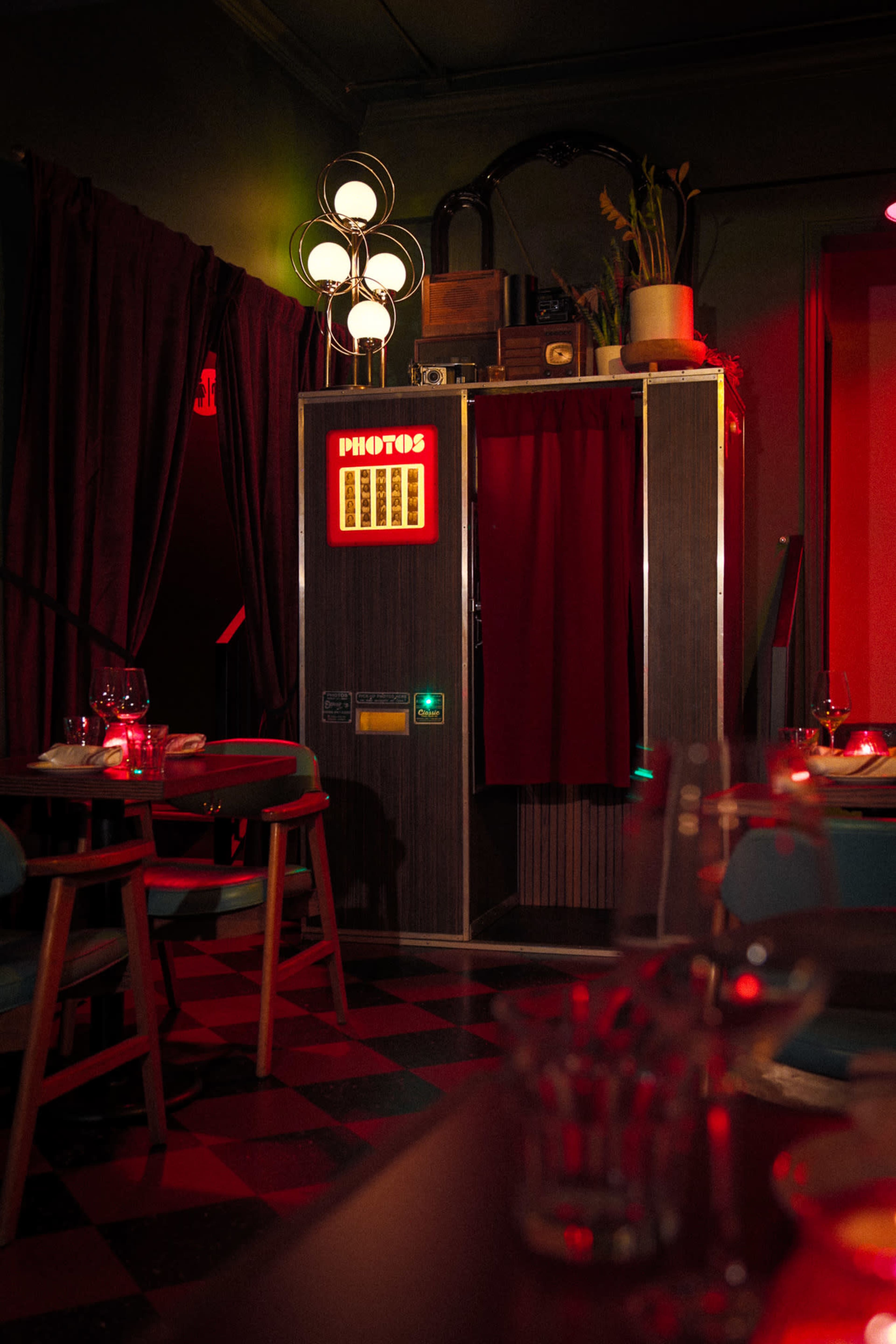 A vintage-style photo booth with a red curtain is located in a dimly lit restaurant, surrounded by tables and chairs.