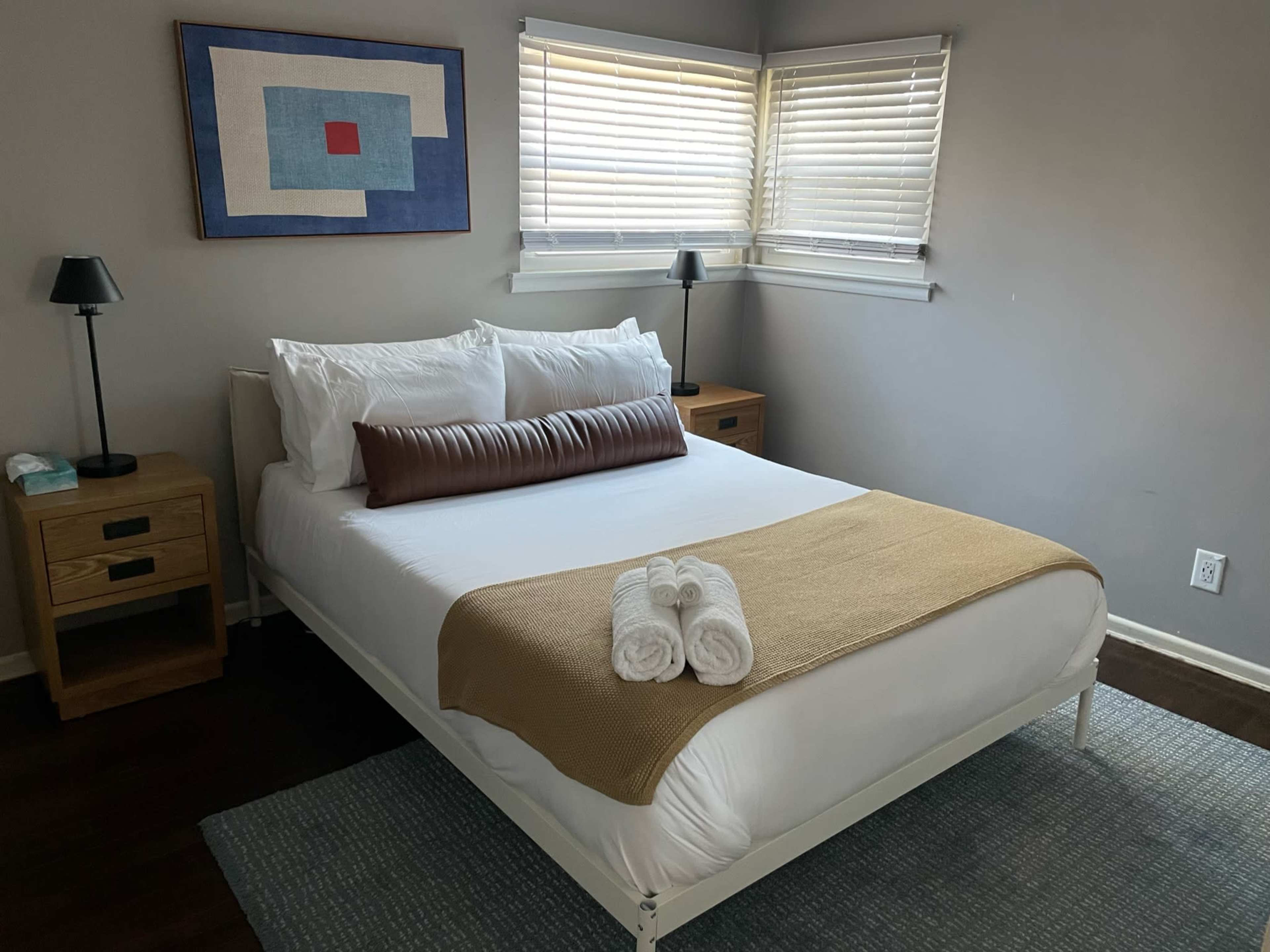 A neatly arranged bedroom featuring a bed with white linens, a brown cushion, a beige throw blanket, two bedside tables with lamps, and a window with blinds.