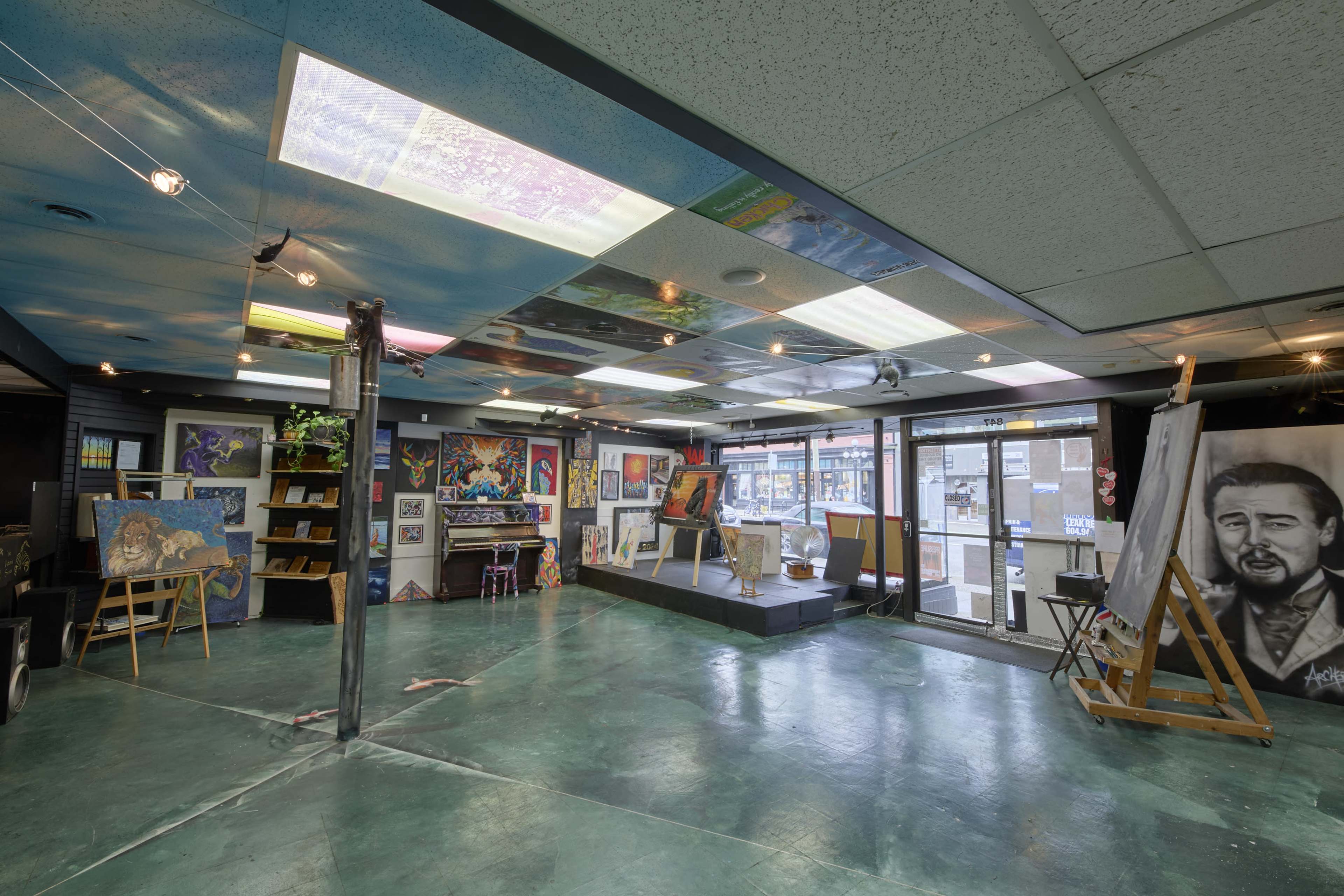 The image shows an art gallery interior with colorful artworks displayed on walls and various art supplies arranged around the space.