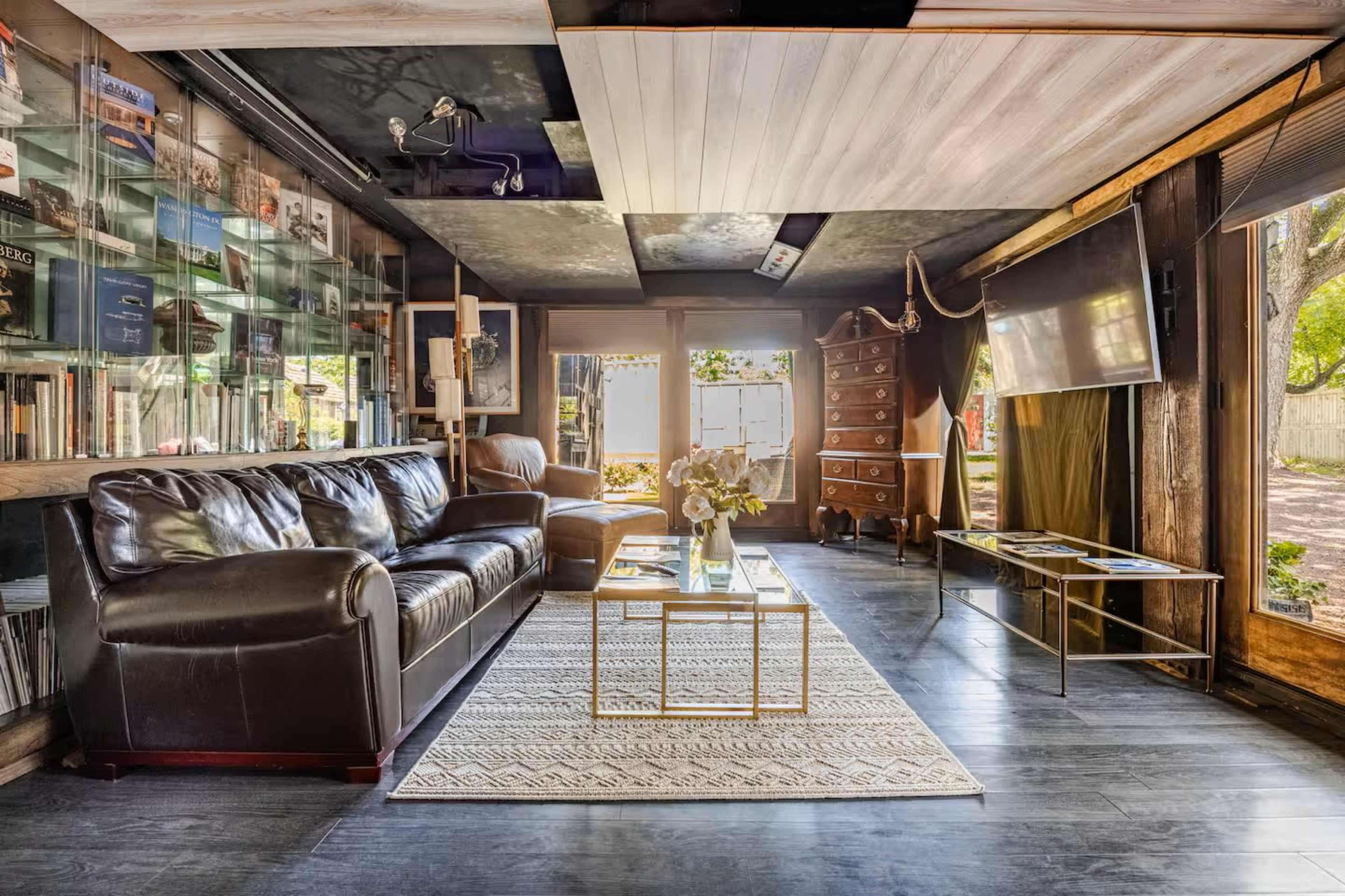 The image shows a modern living room featuring a brown leather couch, a coffee table, and a decorative rug, with a wall of bookshelves and large windows providing natural light.