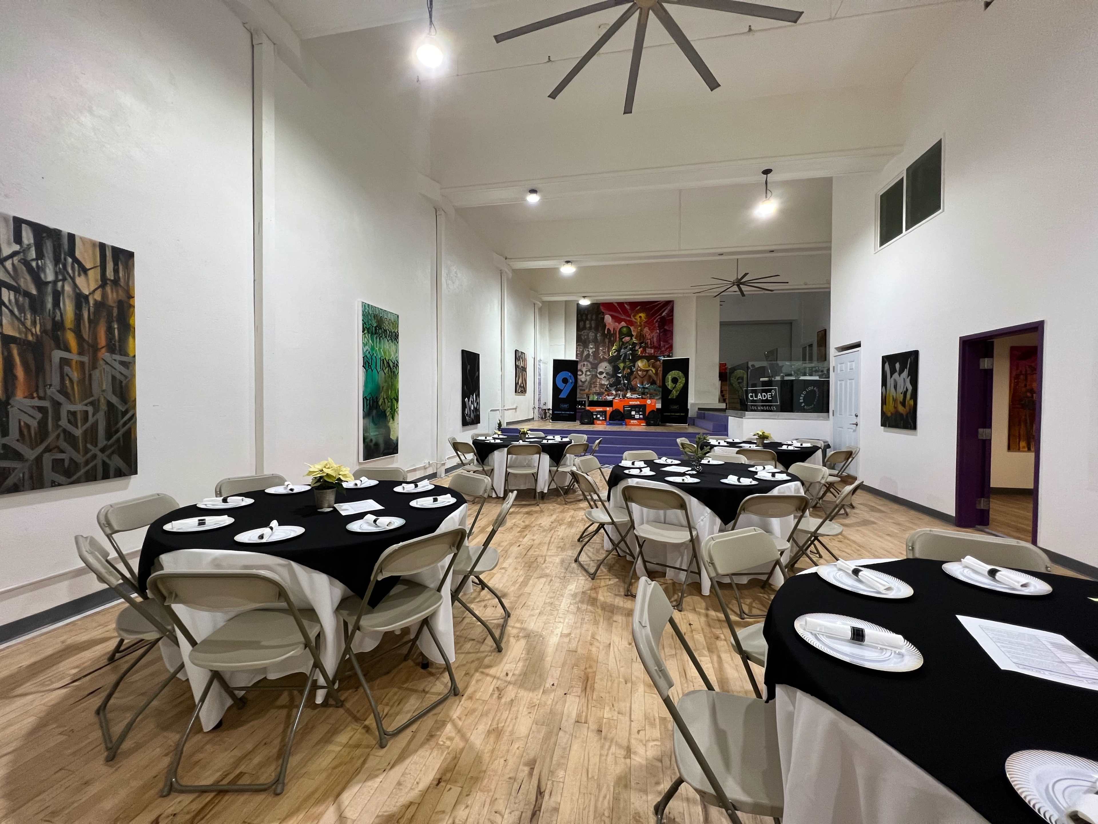 A spacious event room features round tables set with plates and chairs, adorned with artwork on the walls and a large mural in the background.