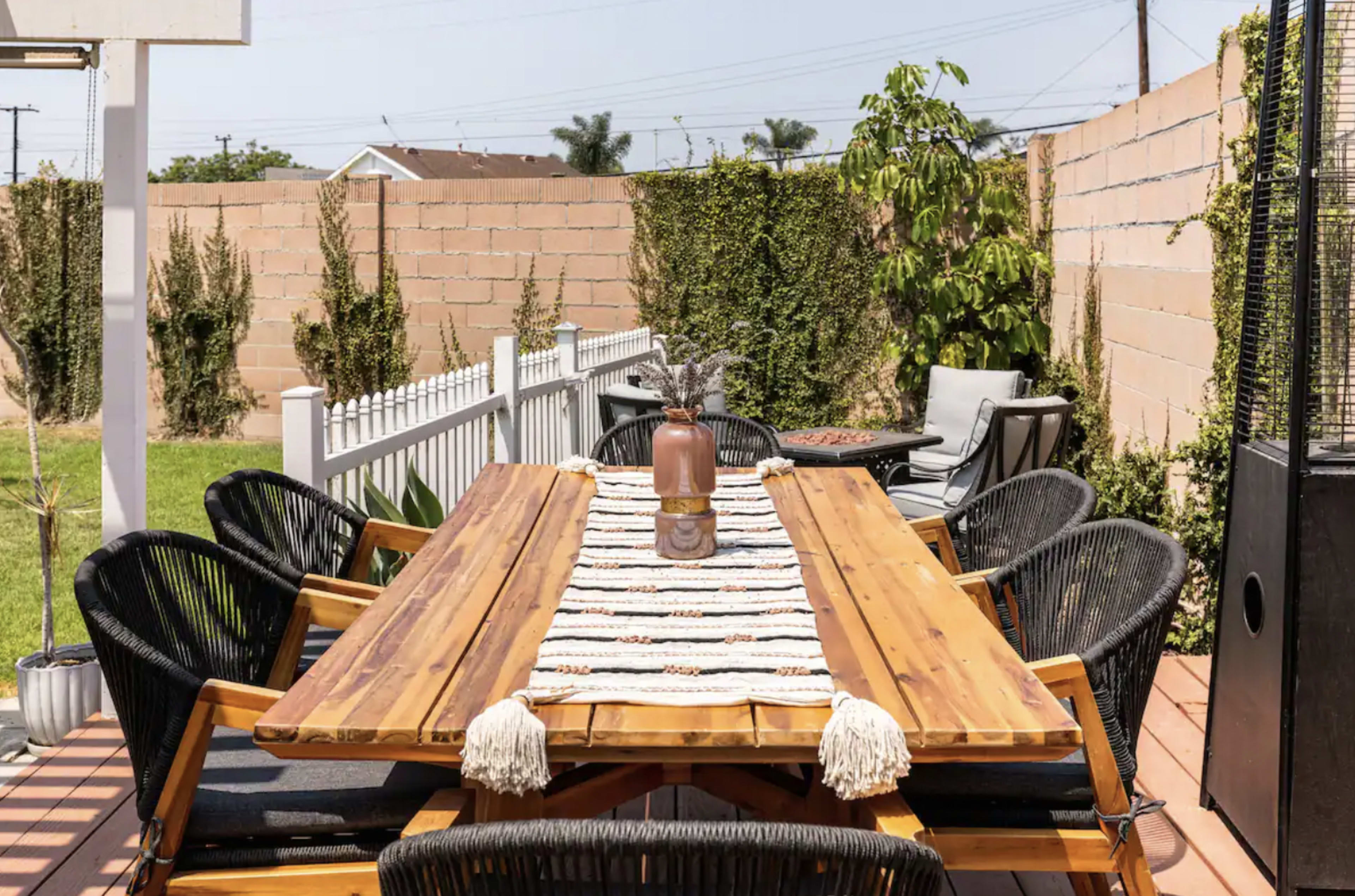 A large wooden dining table is set outdoors on a patio surrounded by black chairs and bordered by a green lawn and brick walls.